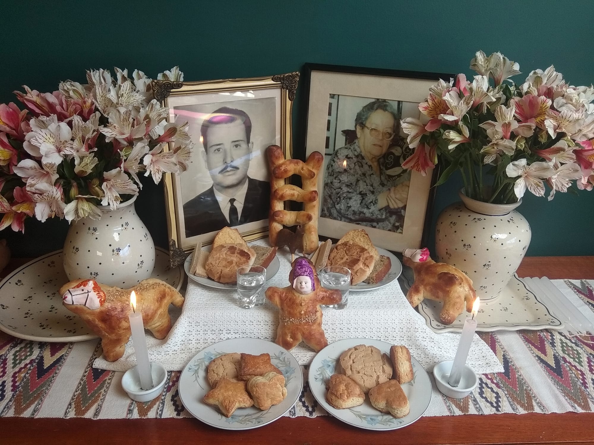 A table set against a green wall with two framed black-and-white portraits, vases of pink and white lilies, a ladder made of bread, a bread figure with a painted face, bread horses, plates of biscuits, glasses of water and two lit candles on a woven runner