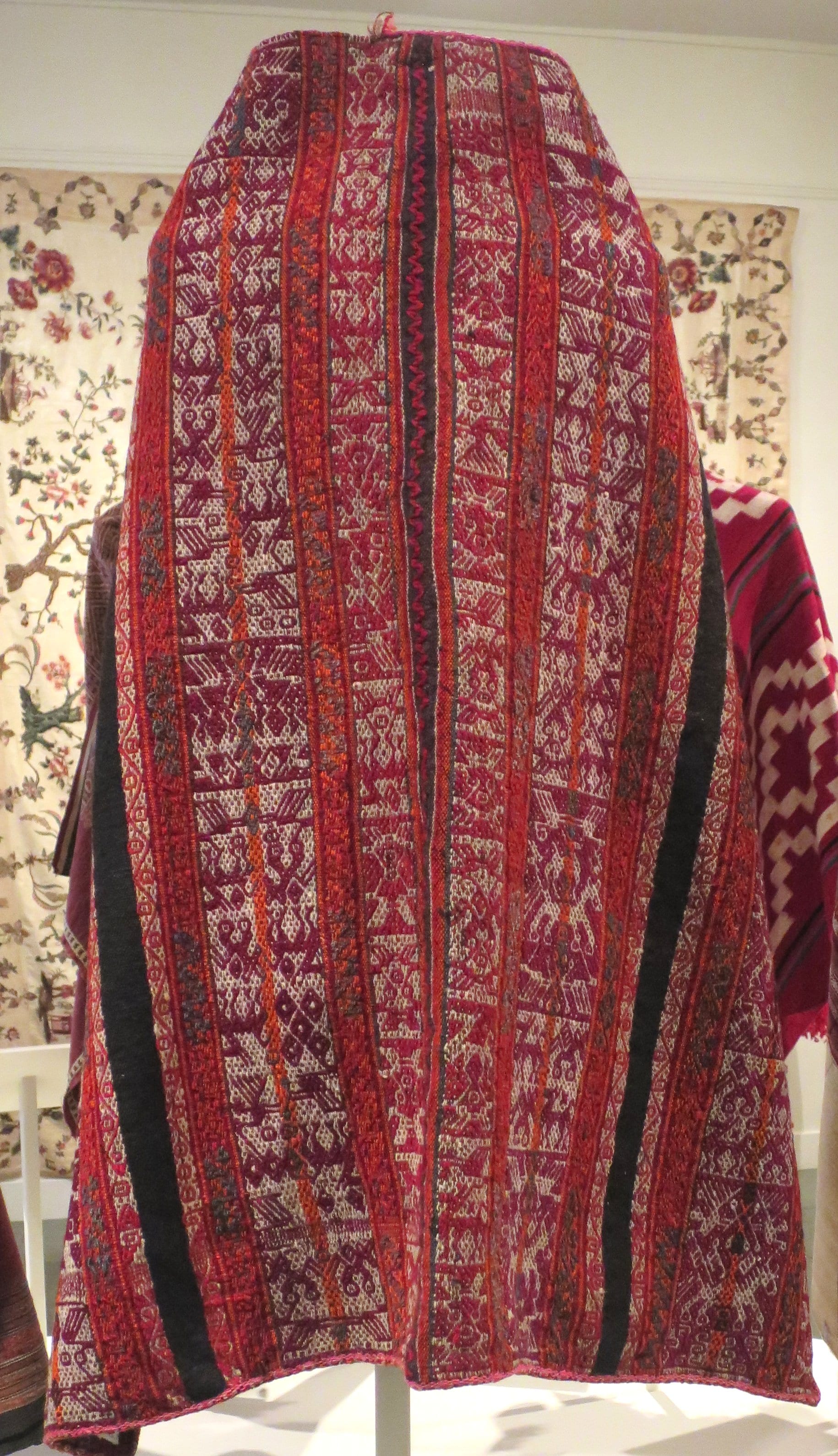 A handwoven wool mantle displayed on a stand, with narrow vertical bands of red, magenta, cream and black filled with fine geometric and figurative patterns