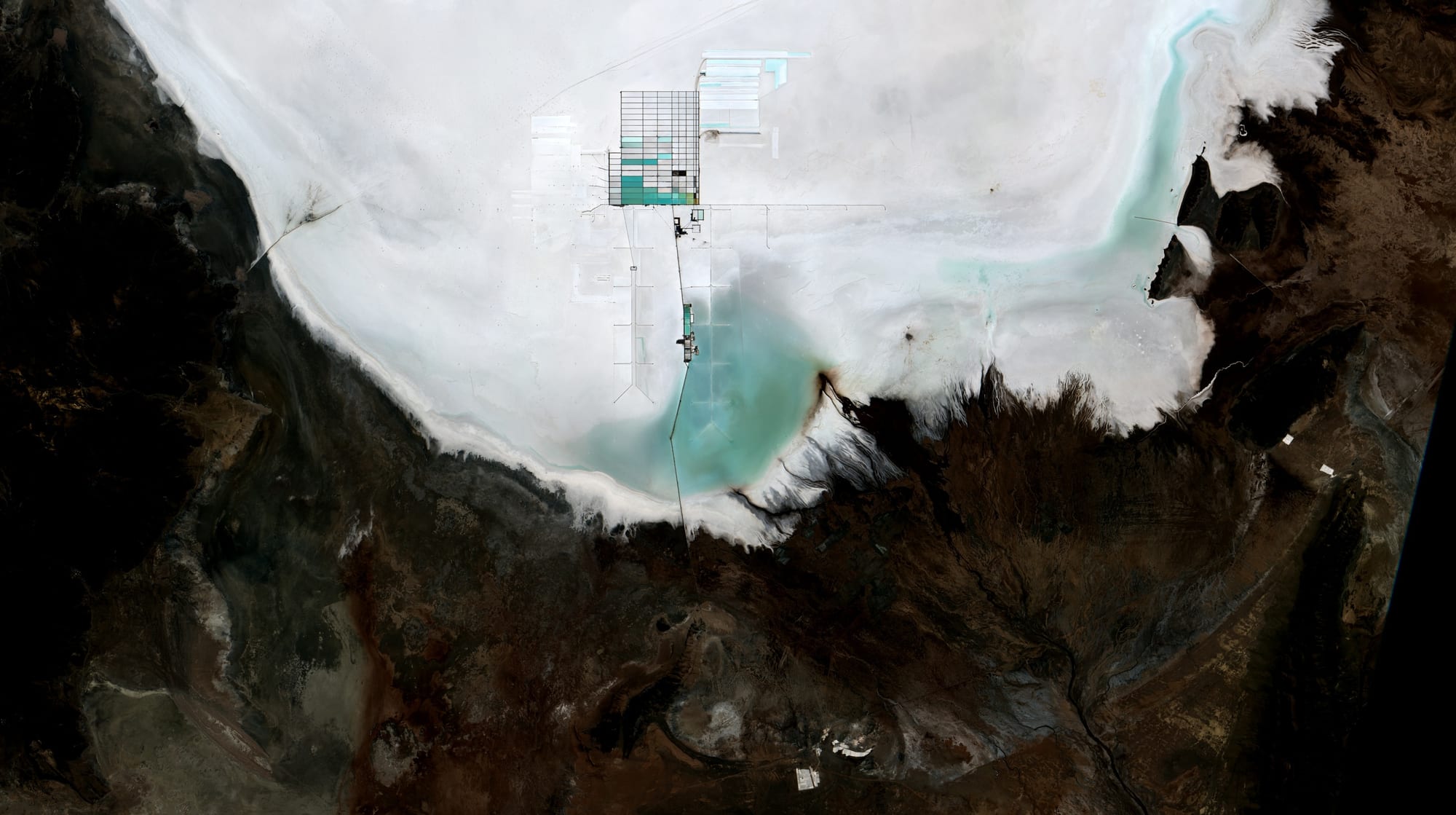 A satellite view of the white southern edge of a salt flat against dark brown land, with a chequered grid of turquoise ponds set into the salt