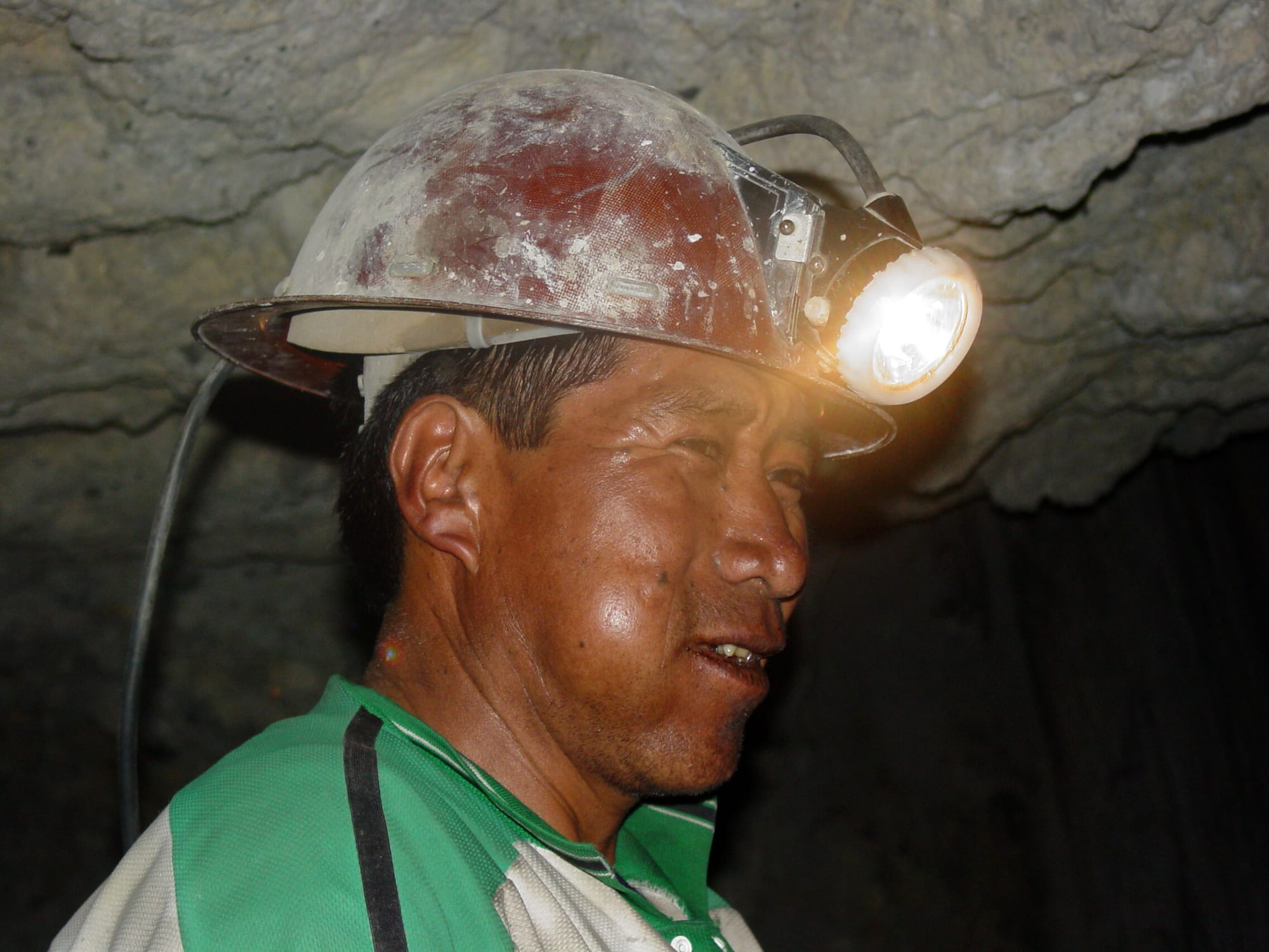 A miner in a hard hat crouched in a low, rough tunnel lit by his headlamp