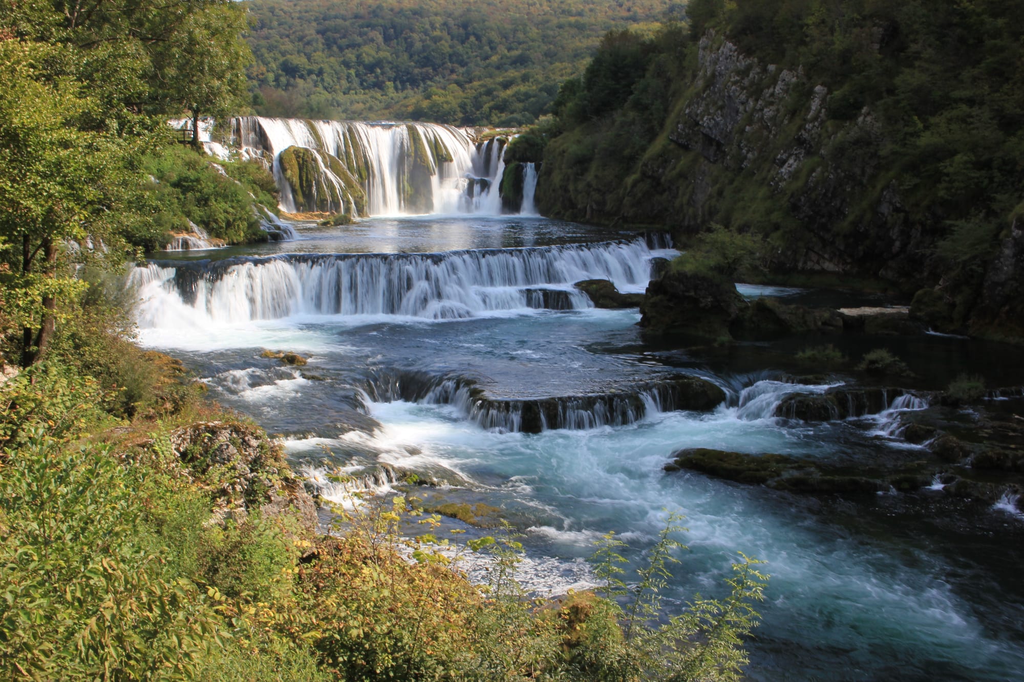 A powerful wide waterfall stepping over travertine ledges in a forested river canyon