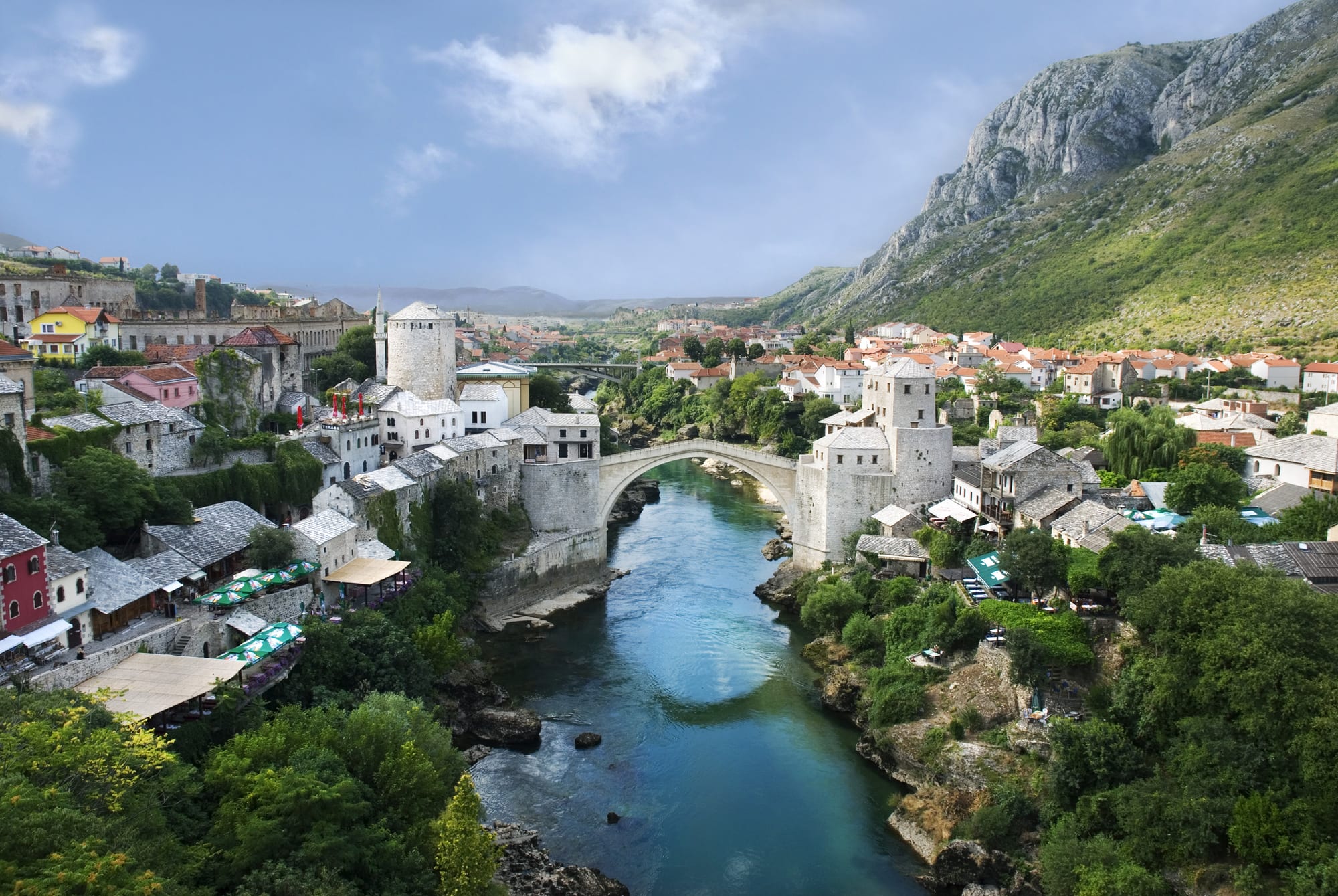 A high single-arched stone bridge over an emerald river, minarets and stone houses on both banks