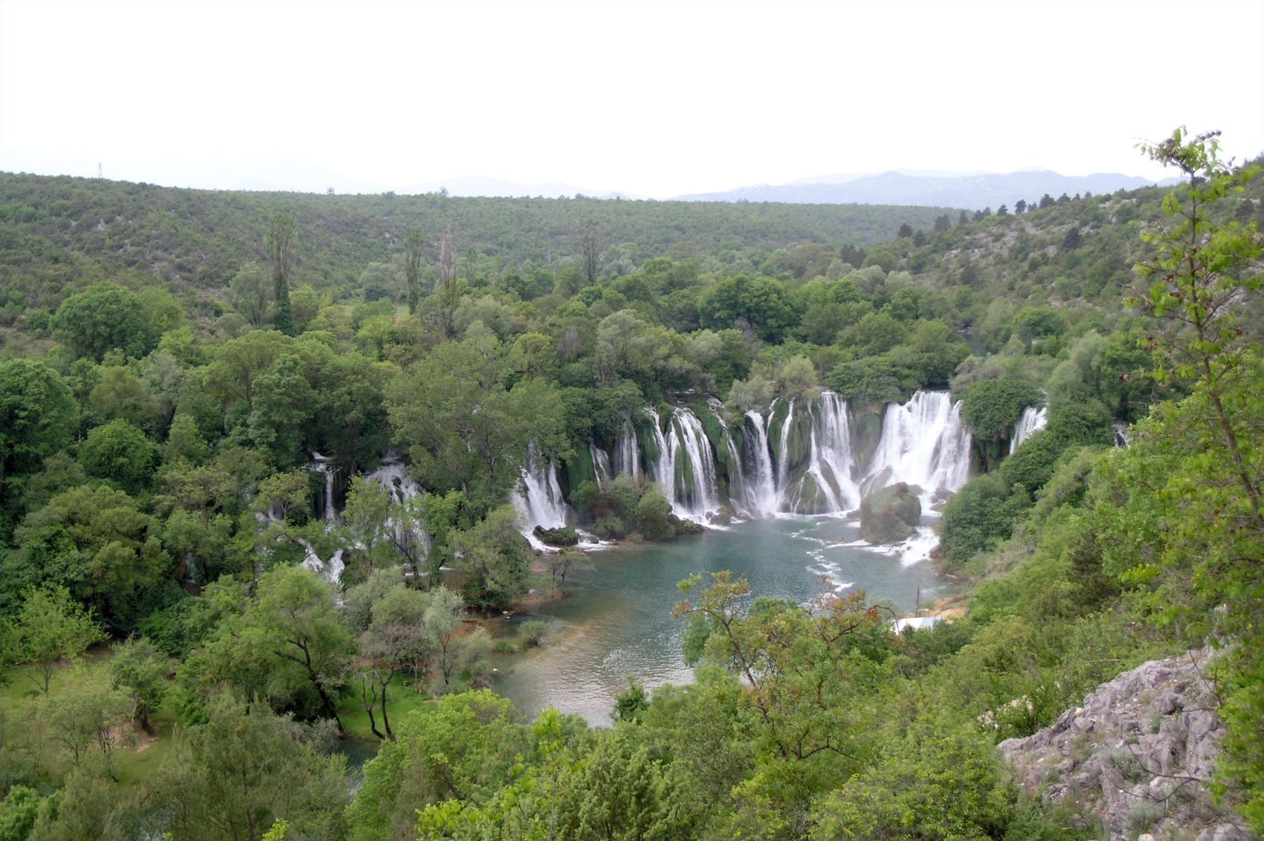 A wide semicircle of waterfalls dropping into a large green pool ringed by trees
