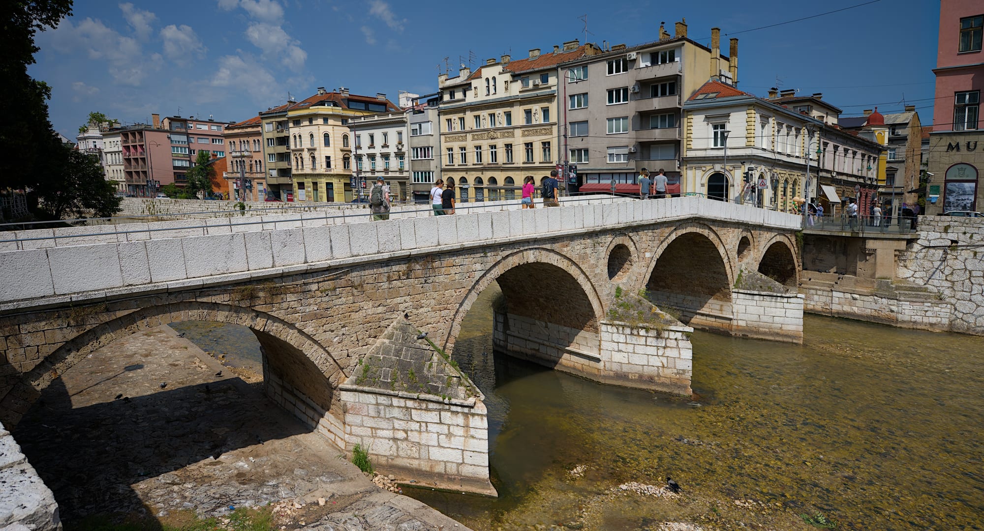 A low Ottoman stone bridge of four arches crossing a shallow river, with a row of Austro-Hungarian apartment buildings on the far bank