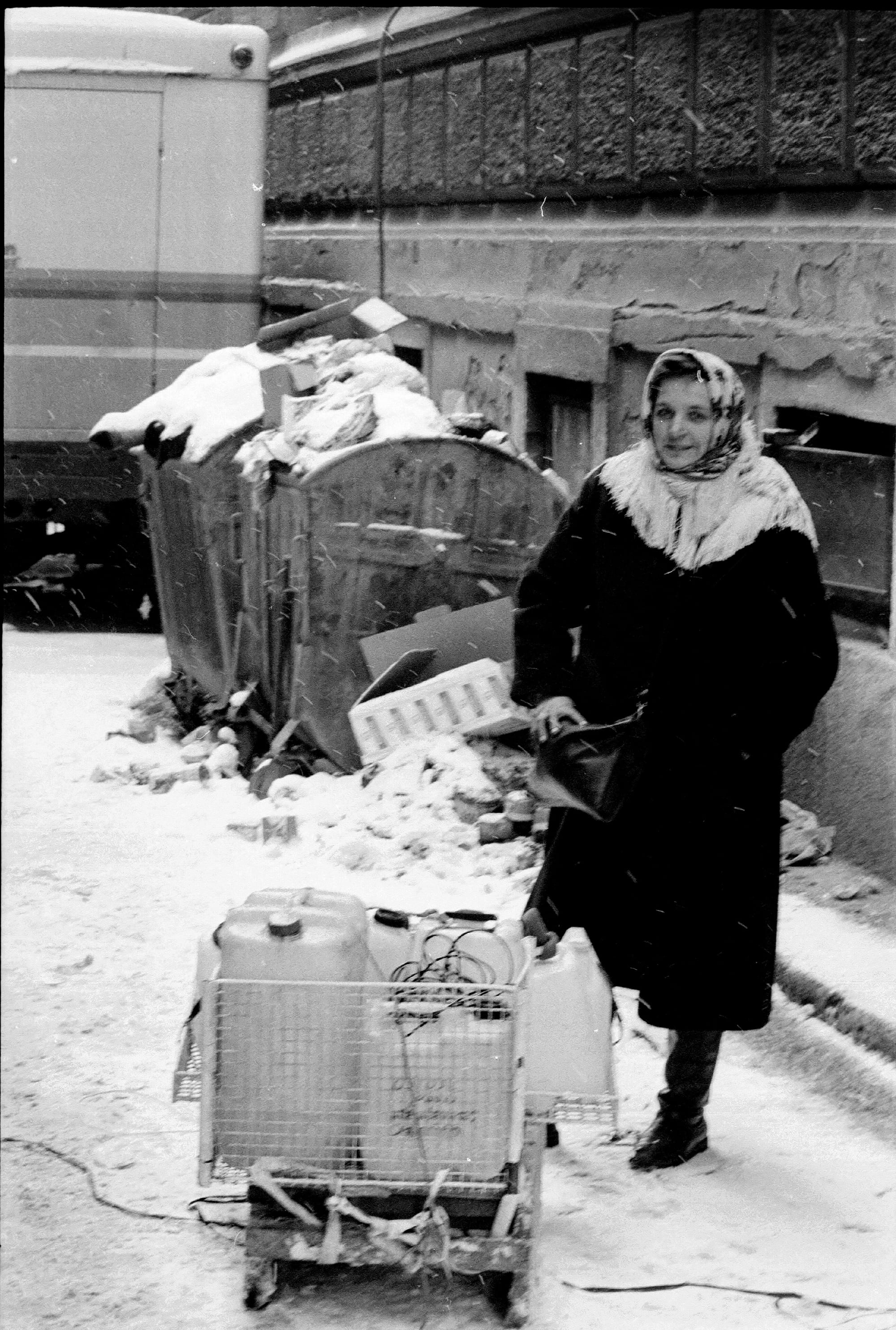 People hurrying with plastic containers along a damaged street past a wall pocked with shell craters