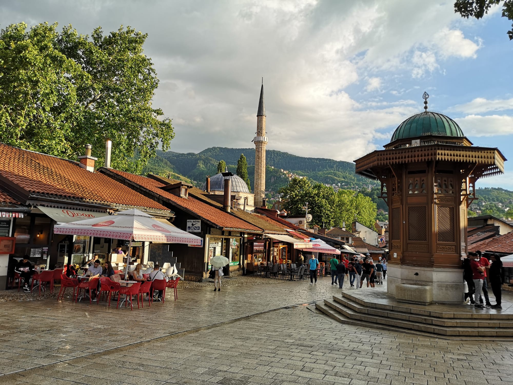 A domed Ottoman fountain kiosk in a cobbled square ringed by low shops, minarets and hills behind
