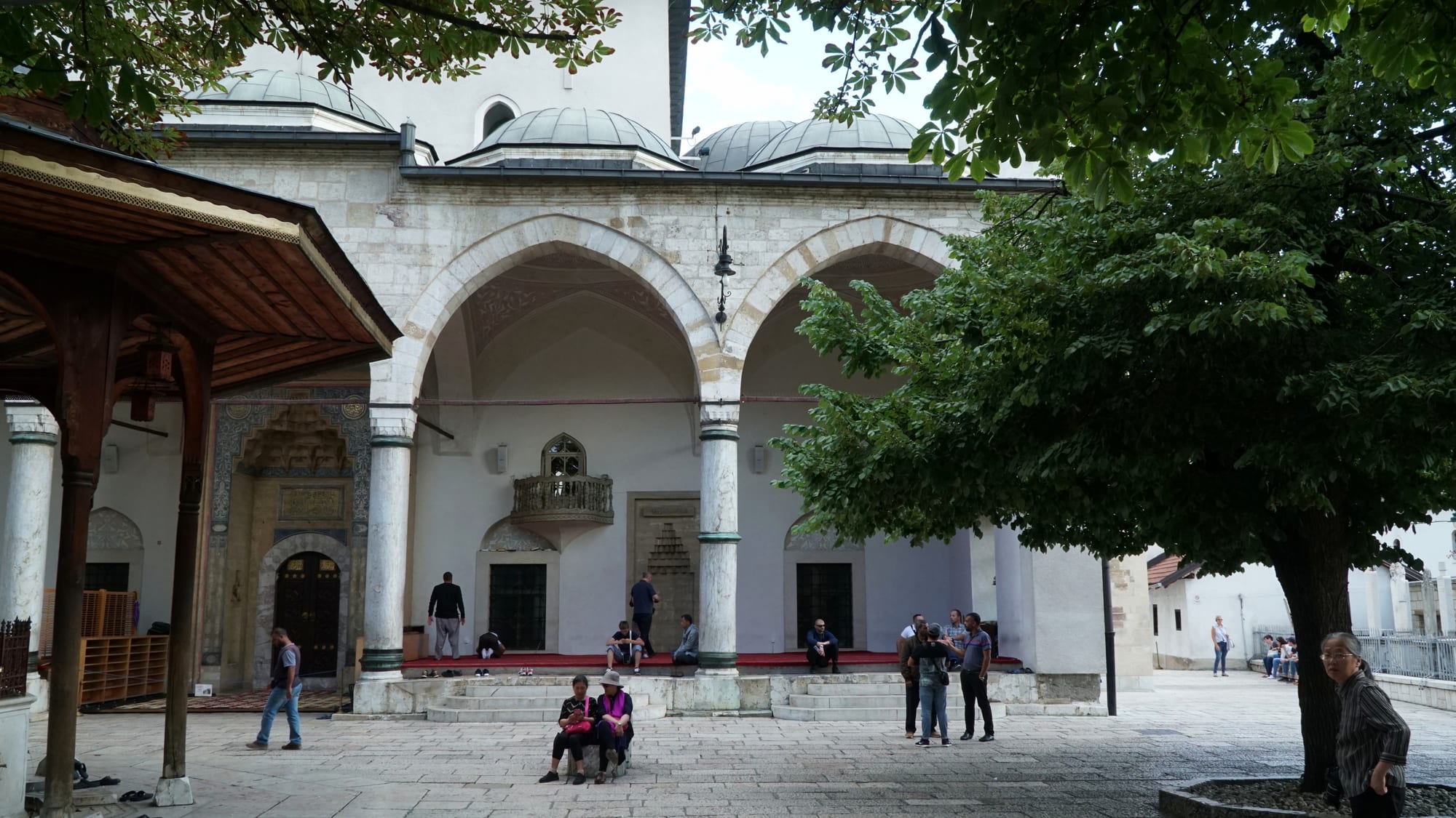 The arched stone portico of a domed Ottoman mosque, with people standing and sitting in a paved courtyard shaded by a large tree