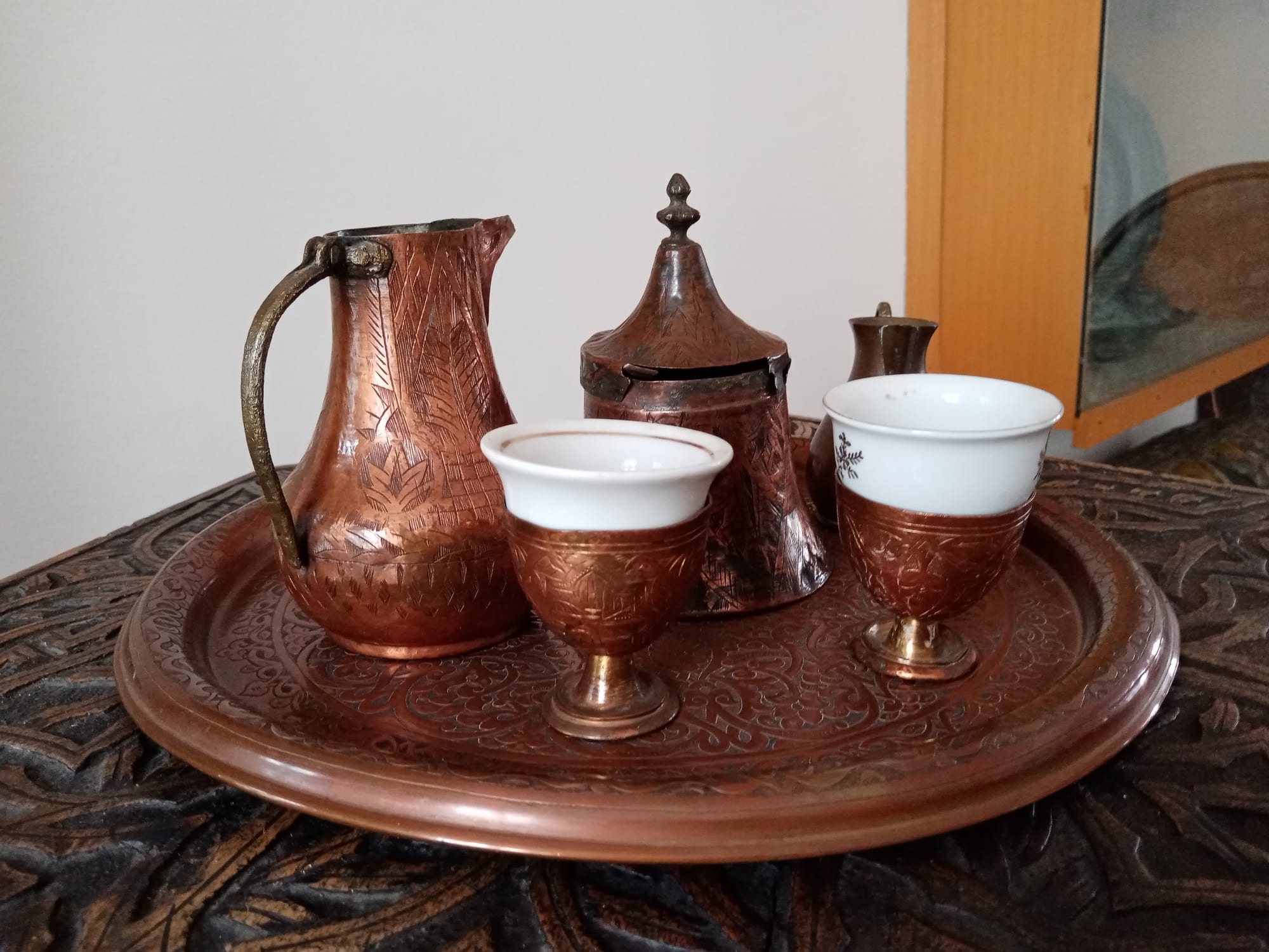 An engraved copper tray on a carved wooden table holding a tall copper jug, a lidded copper pot and two small white cups set in copper holders