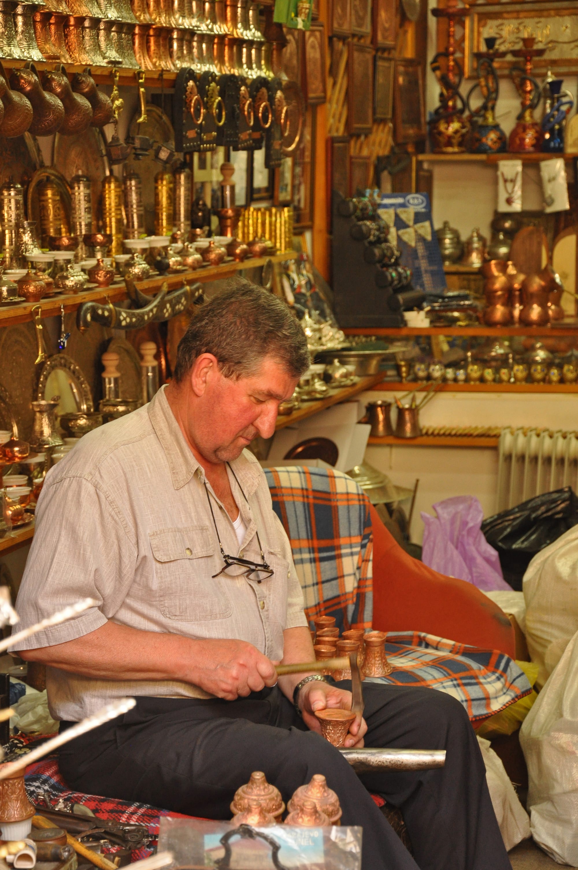 A seated craftsman taps a pattern into a small copper vessel with a hammer, surrounded by shelves of copper coffee pots, trays and ornaments