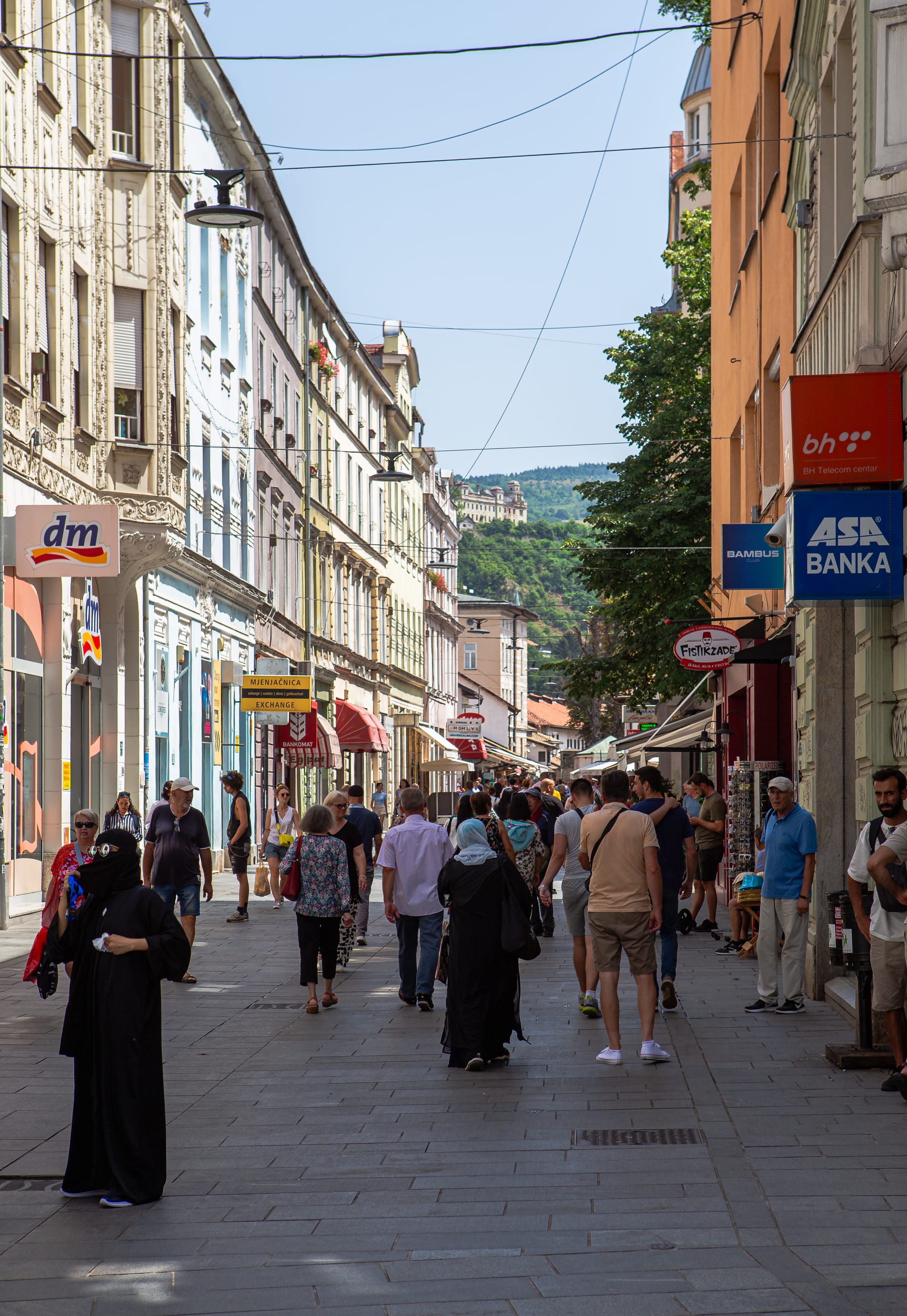 A crowded pedestrian shopping street in Sarajevo lined with shops and banks, wooded hills visible at the far end
