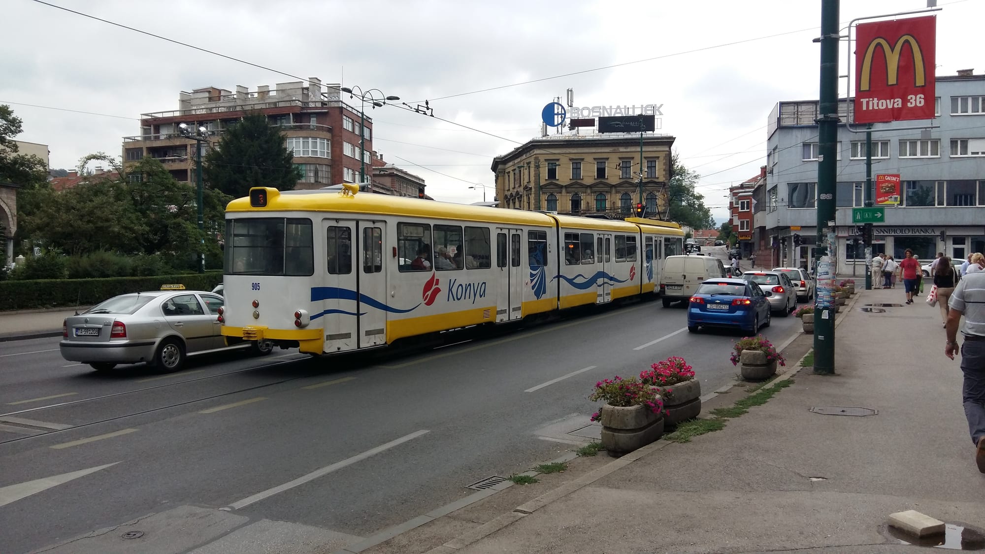 A yellow and white tram running along a Sarajevo street lined with apartment blocks, cars queued alongside it