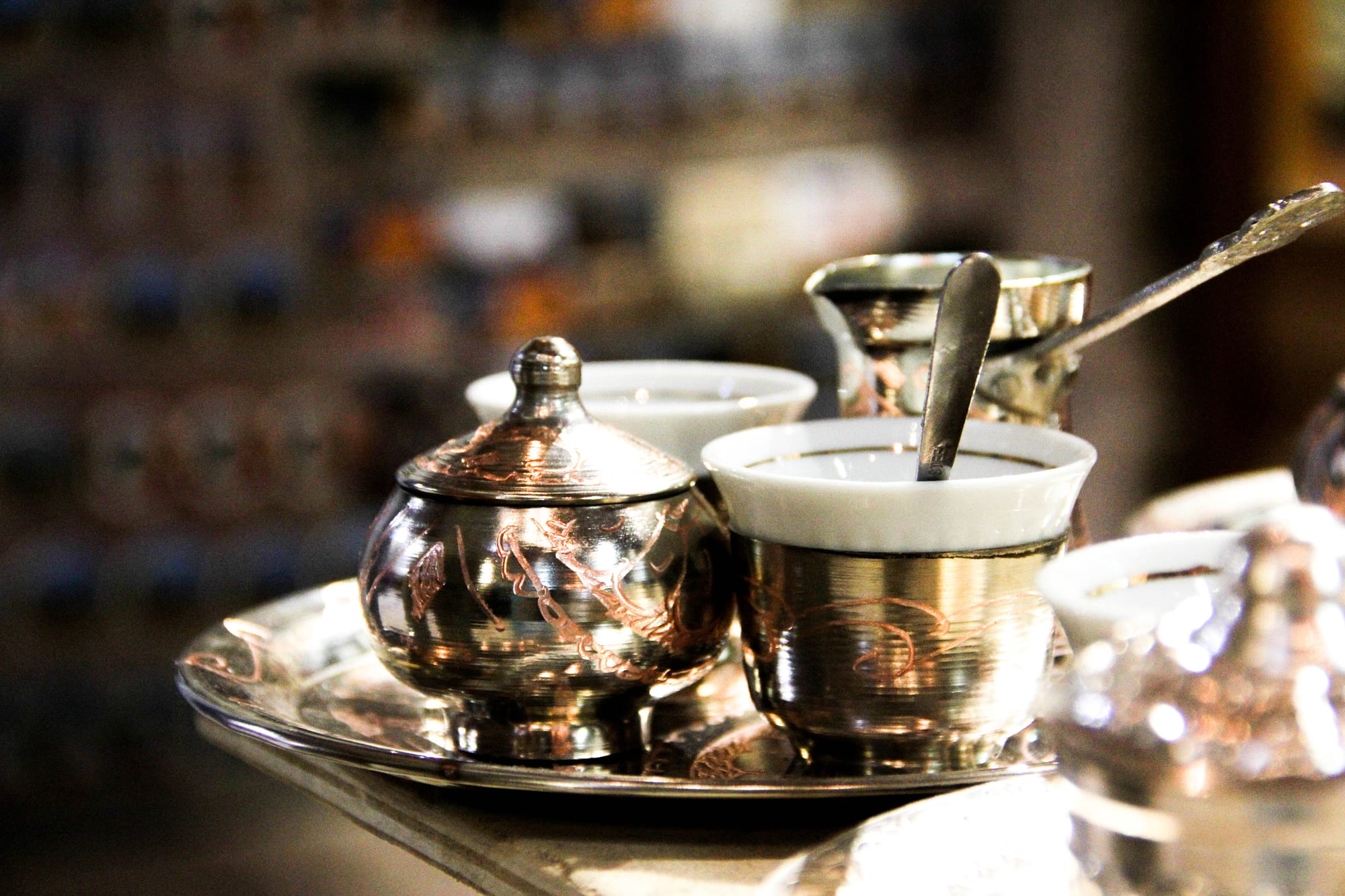 A close-up of a small white cup of Bosnian coffee with a spoon, set on a metal tray beside a copper džezva and sugar bowl