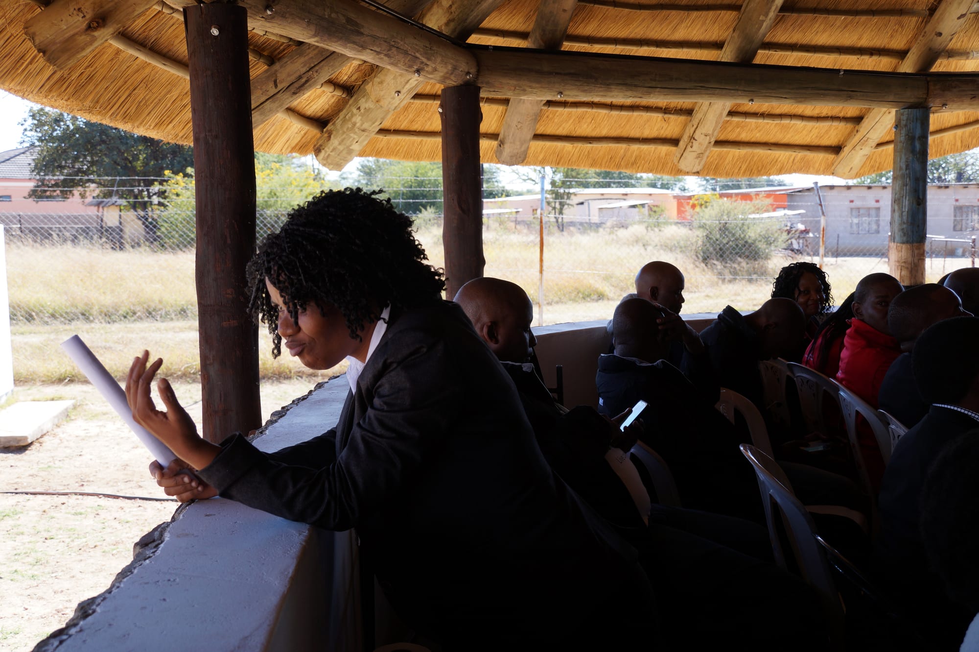 People seated under a large thatched shelter at an outdoor community meeting, with grassland and houses visible beyond the open sides