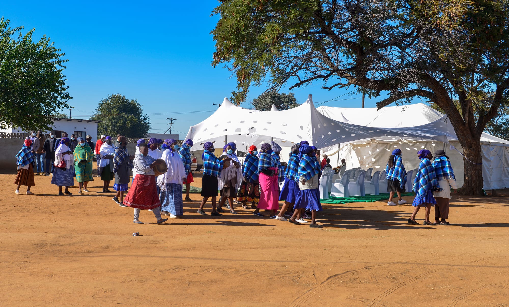 A group of women in blue-checked shawls, headscarves and long skirts walk across a sandy yard in front of a white marquee, with trees and a blue sky behind