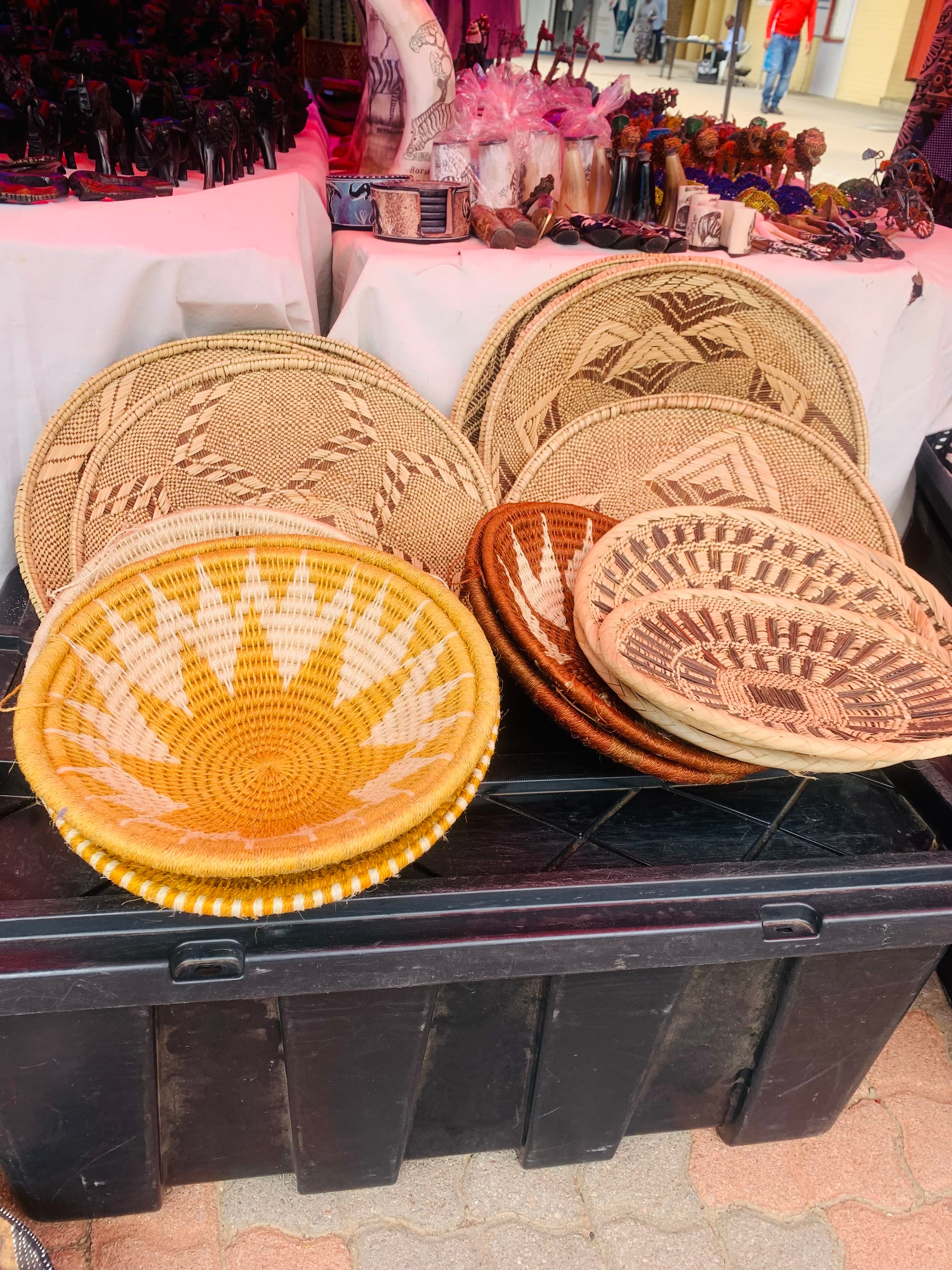 A stall displaying woven baskets and flat trays in natural tan, brown and yellow with geometric and starburst patterns, with carved figures on a table behind