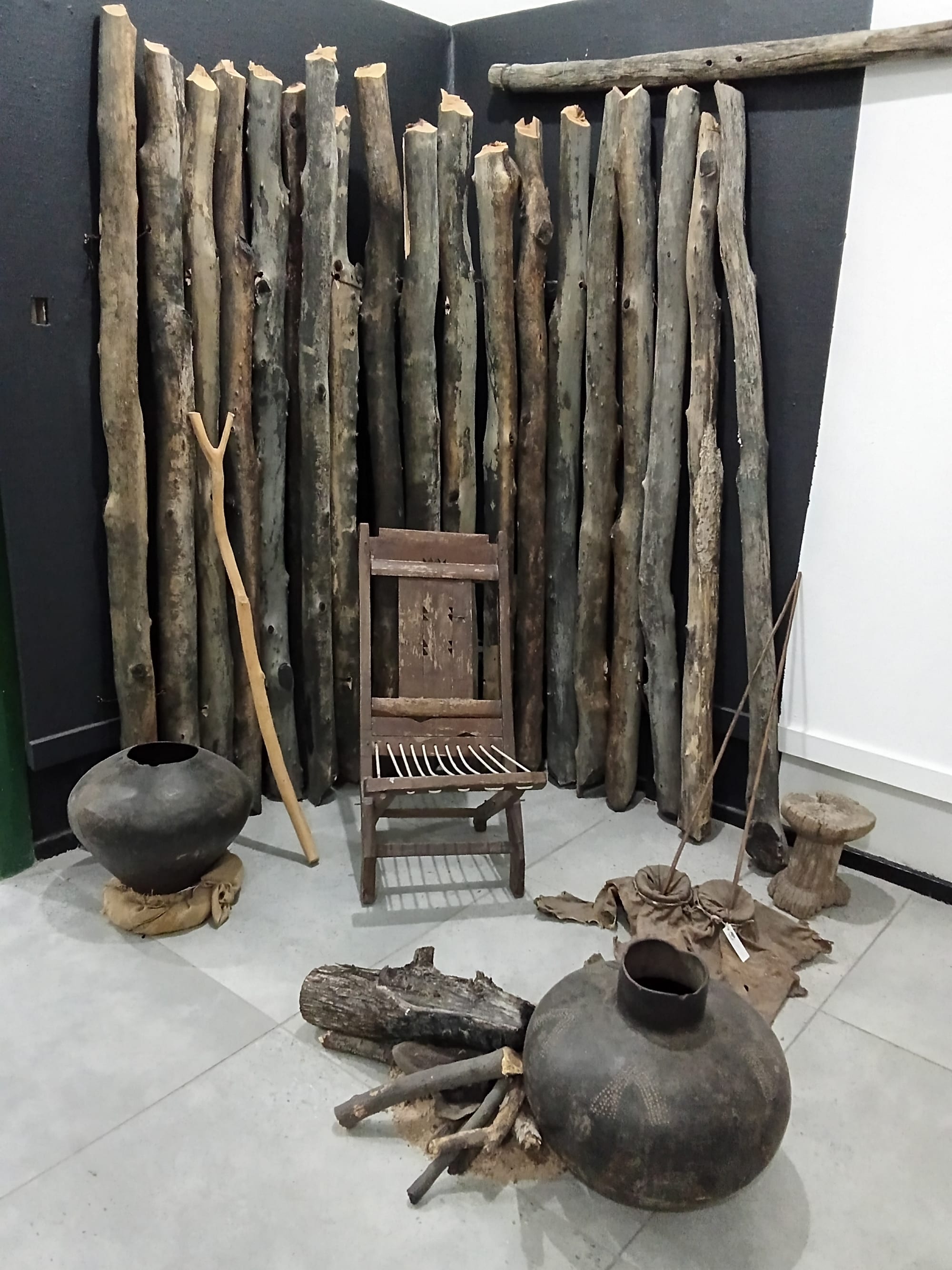 A display recreating a traditional meeting place, with a stockade of upright wooden poles, a wooden folding chair, large dark clay pots and a small pile of firewood