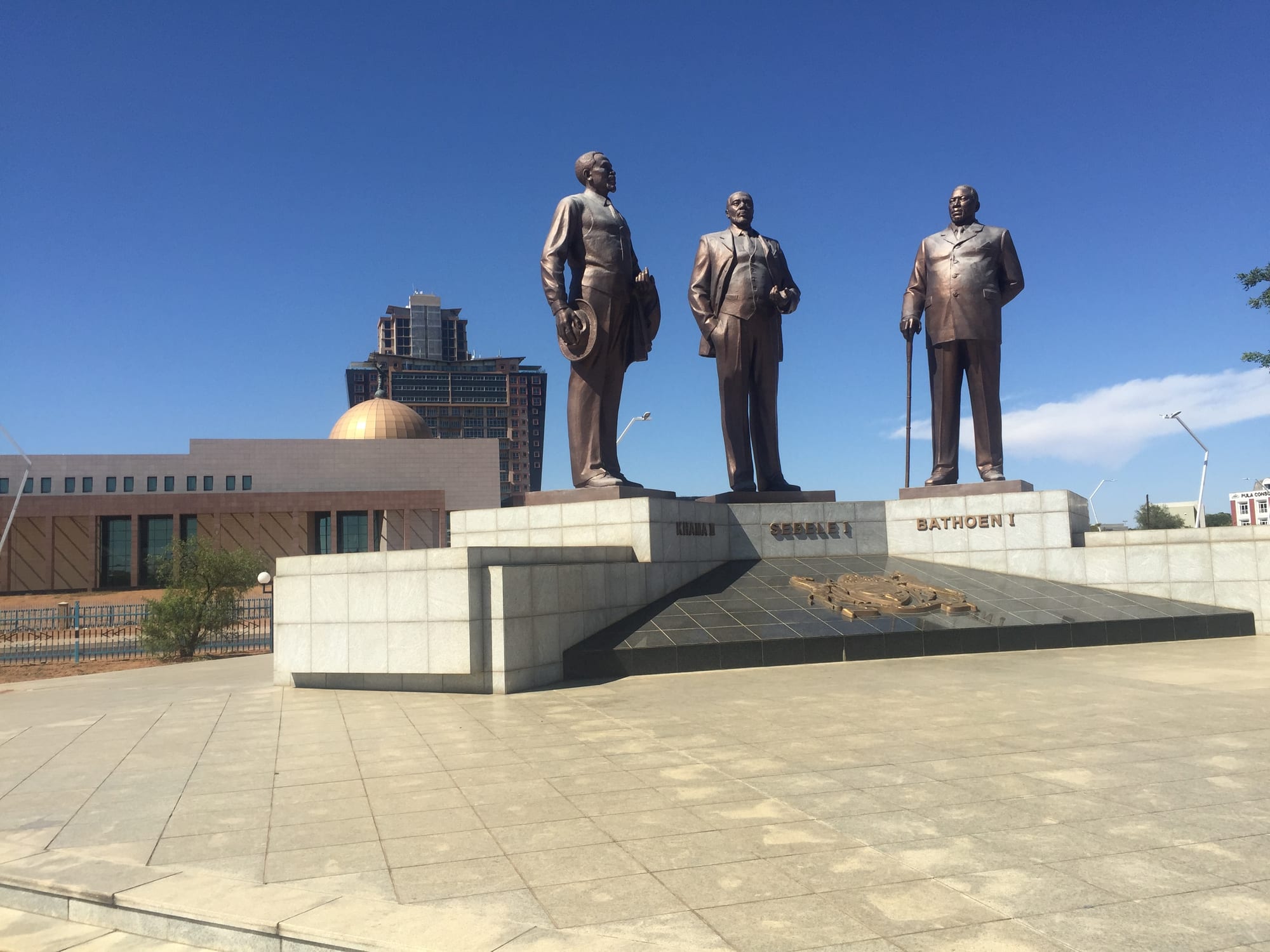 Three tall bronze statues of men in suits standing on a granite plinth in a paved plaza under a deep blue sky, with modern buildings behind