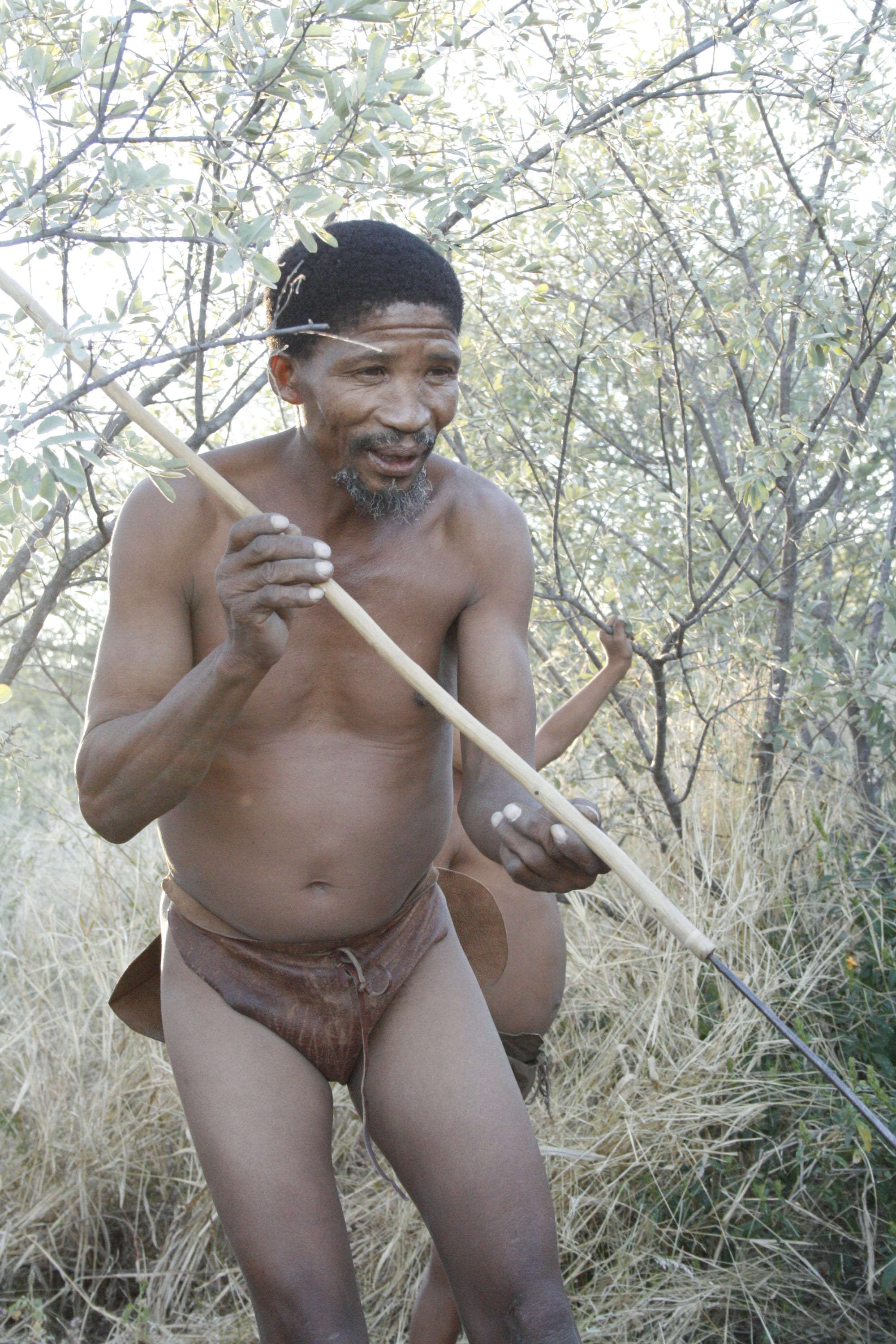 Two San men in light clothing walking with bows and quivers across sparse dry Kalahari grassland