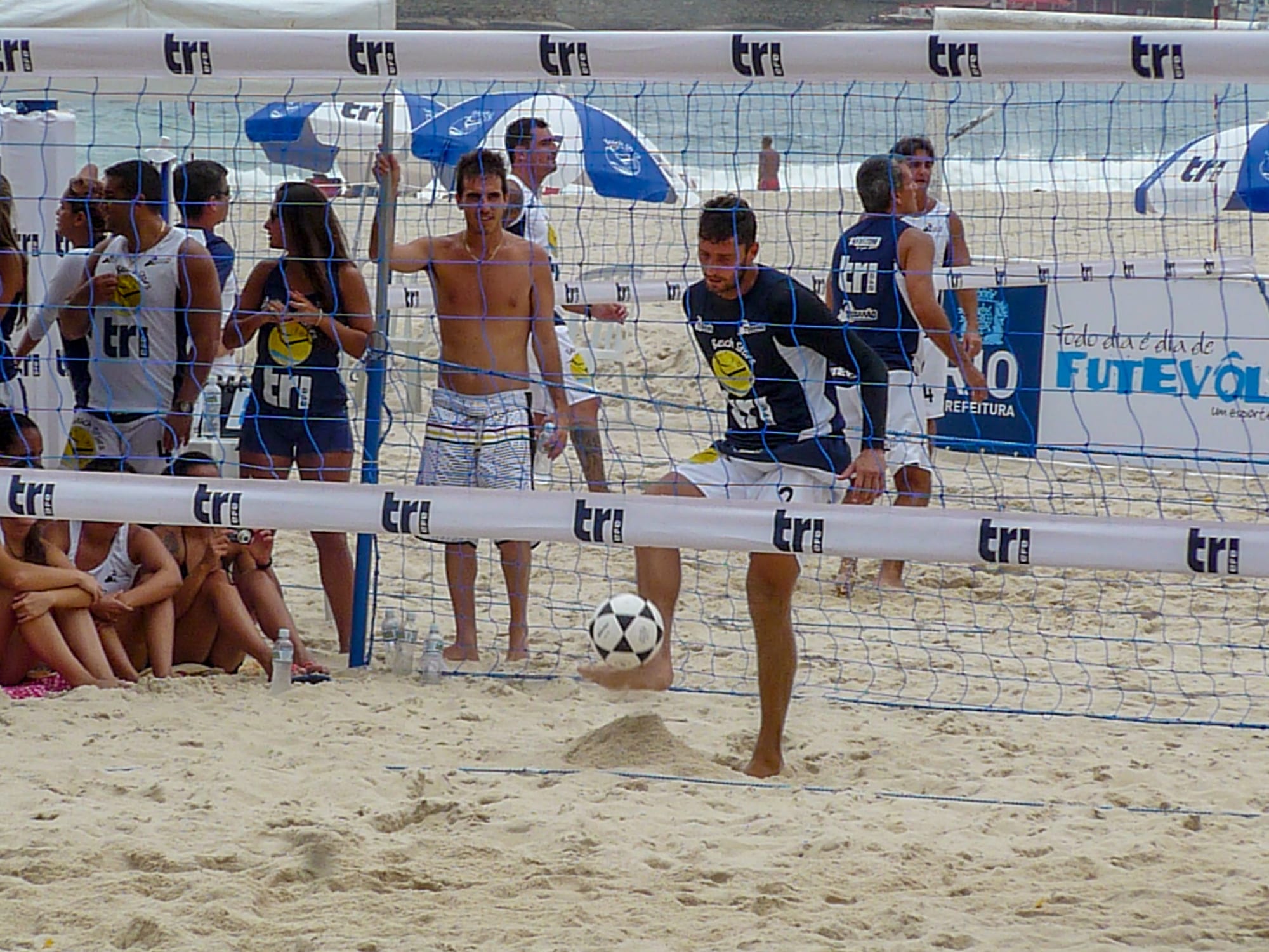 Men playing footvolley, a form of volleyball played with the feet, over a net on a sandy beach with the sea behind