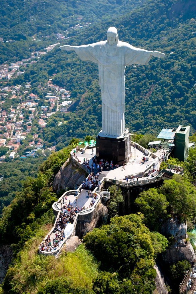 A giant white statue of Christ with arms outstretched on a forested mountain peak above a bay and city