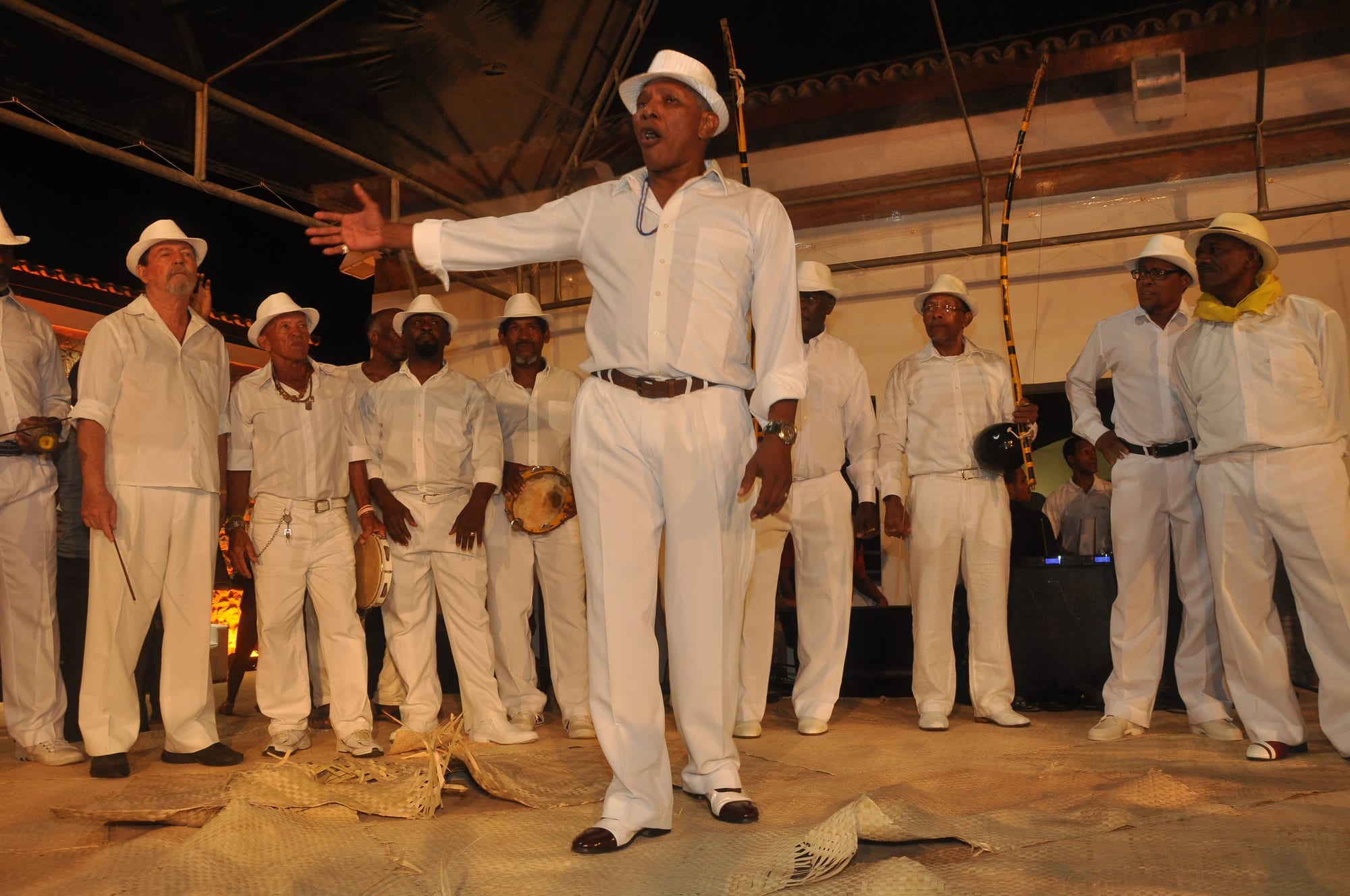 A group of older men dressed in white with white hats stand in a circle, one singing with his arm outstretched, while others hold tambourines and tall bow-shaped berimbaus