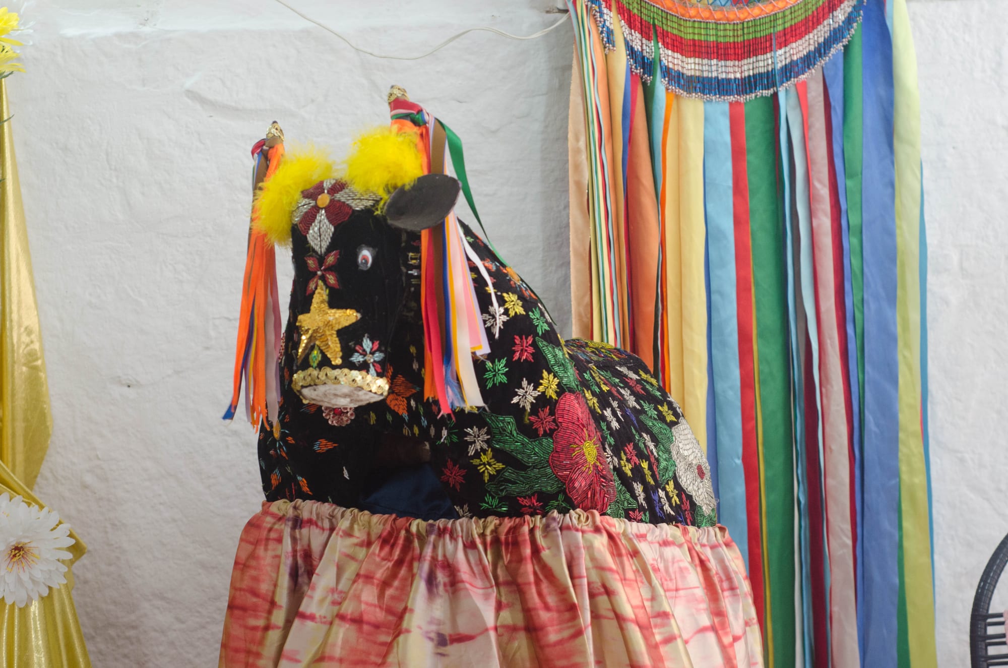A life-size ox figure with a black covering densely embroidered with flowers, stars and beads, hung with coloured ribbons, standing on a pink skirt against a white wall