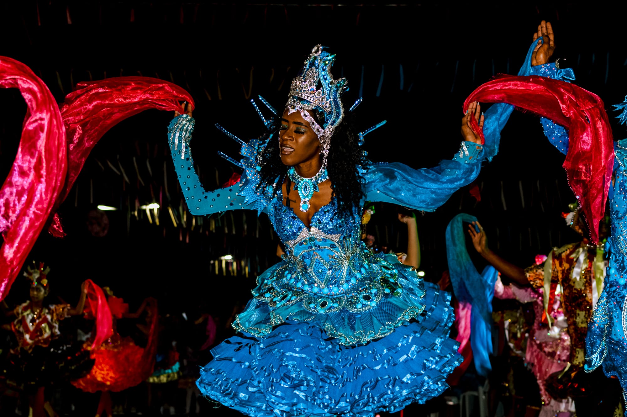 A dancer in a sequinned blue costume and jewelled crown performs with arms outstretched, flanked by dancers waving red and blue fabric