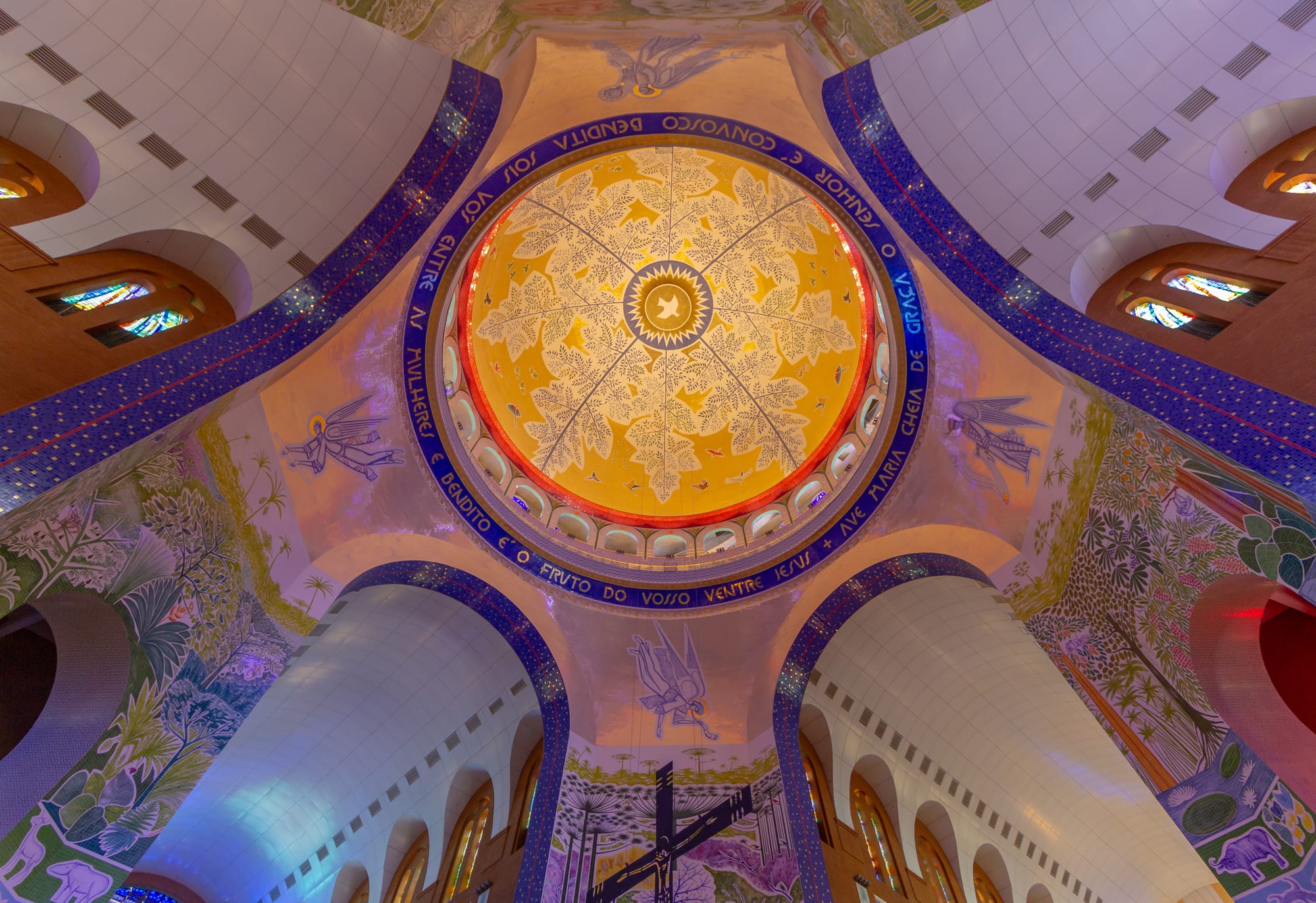 Looking straight up into a church dome painted gold with a leaf pattern, ringed by blue mosaic lettering, with angels and tropical plants painted on the surrounding vaults