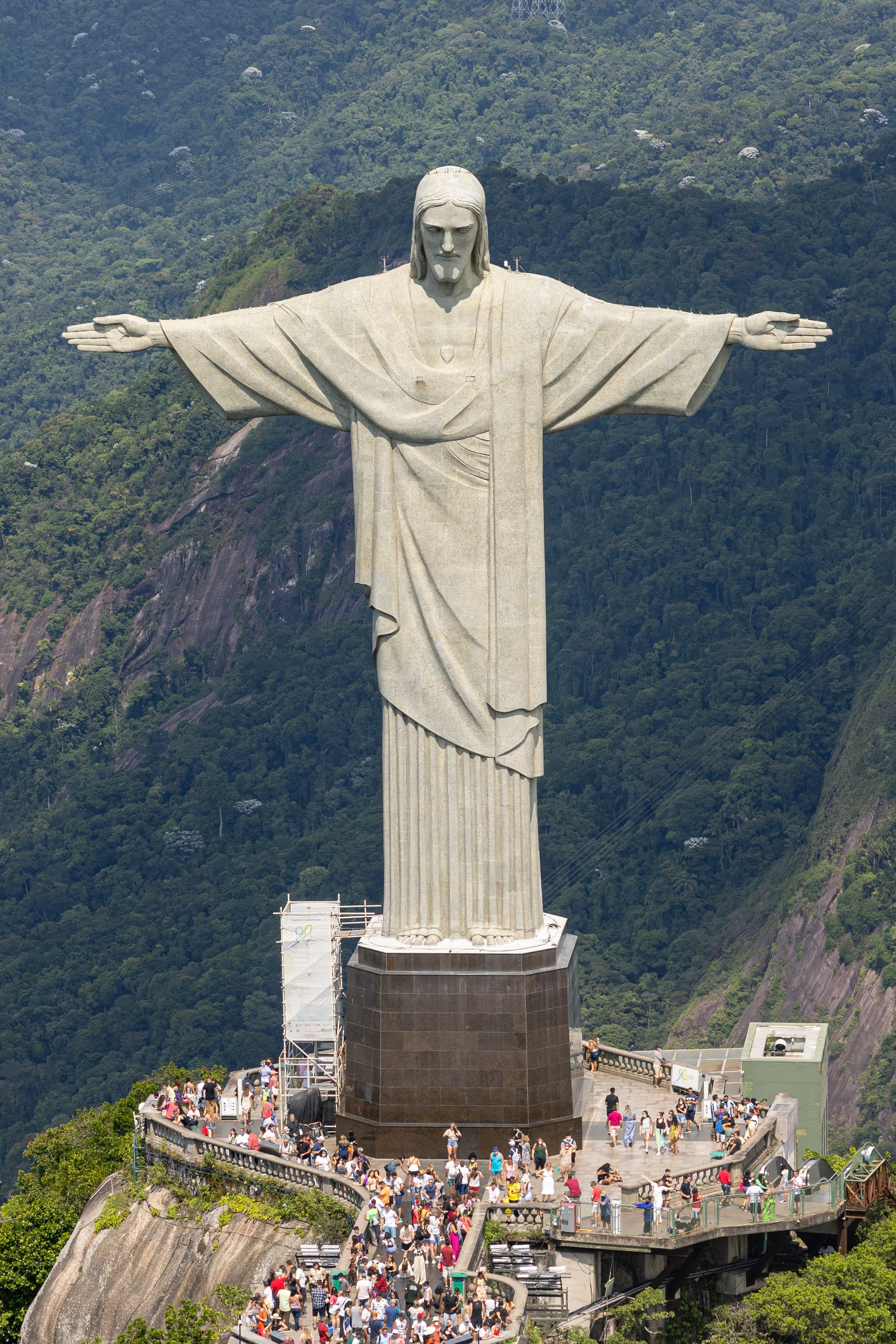 A giant white statue of Christ with outstretched arms on a forested mountain above a bay and city