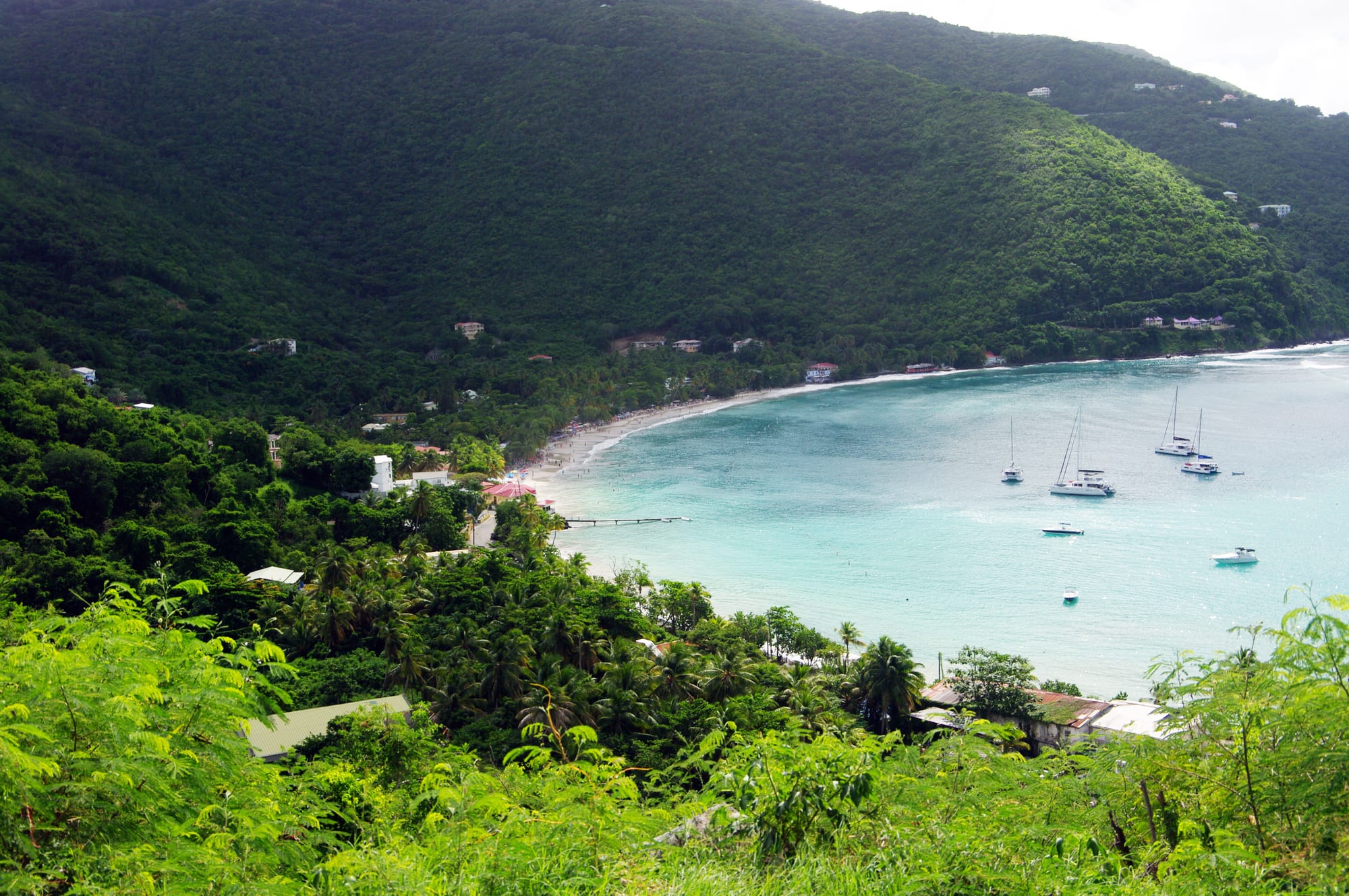 A wide sandy bay backed by green hills and a village, boats at anchor