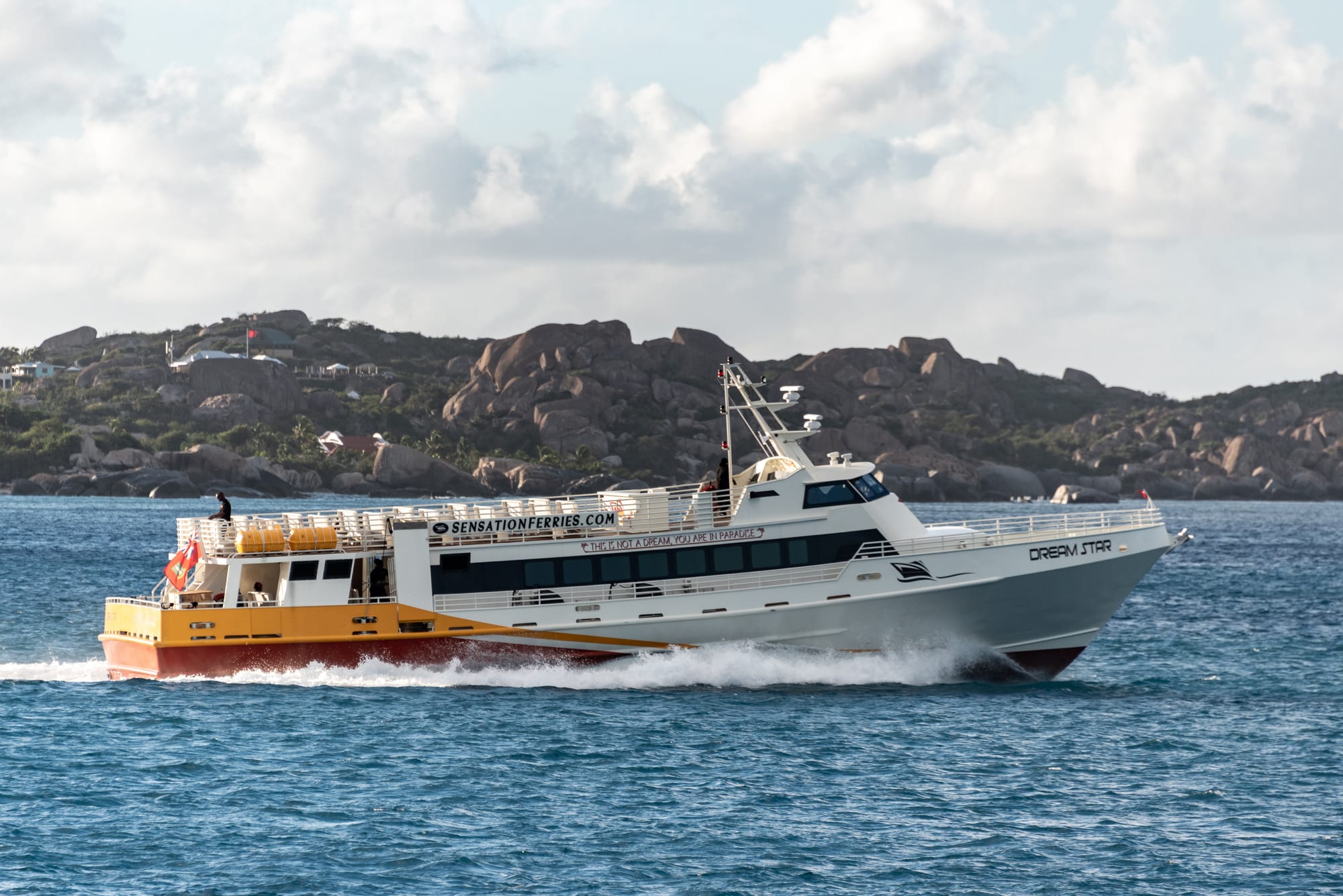 A white and yellow passenger ferry speeding across blue water toward a rocky, hilly coastline