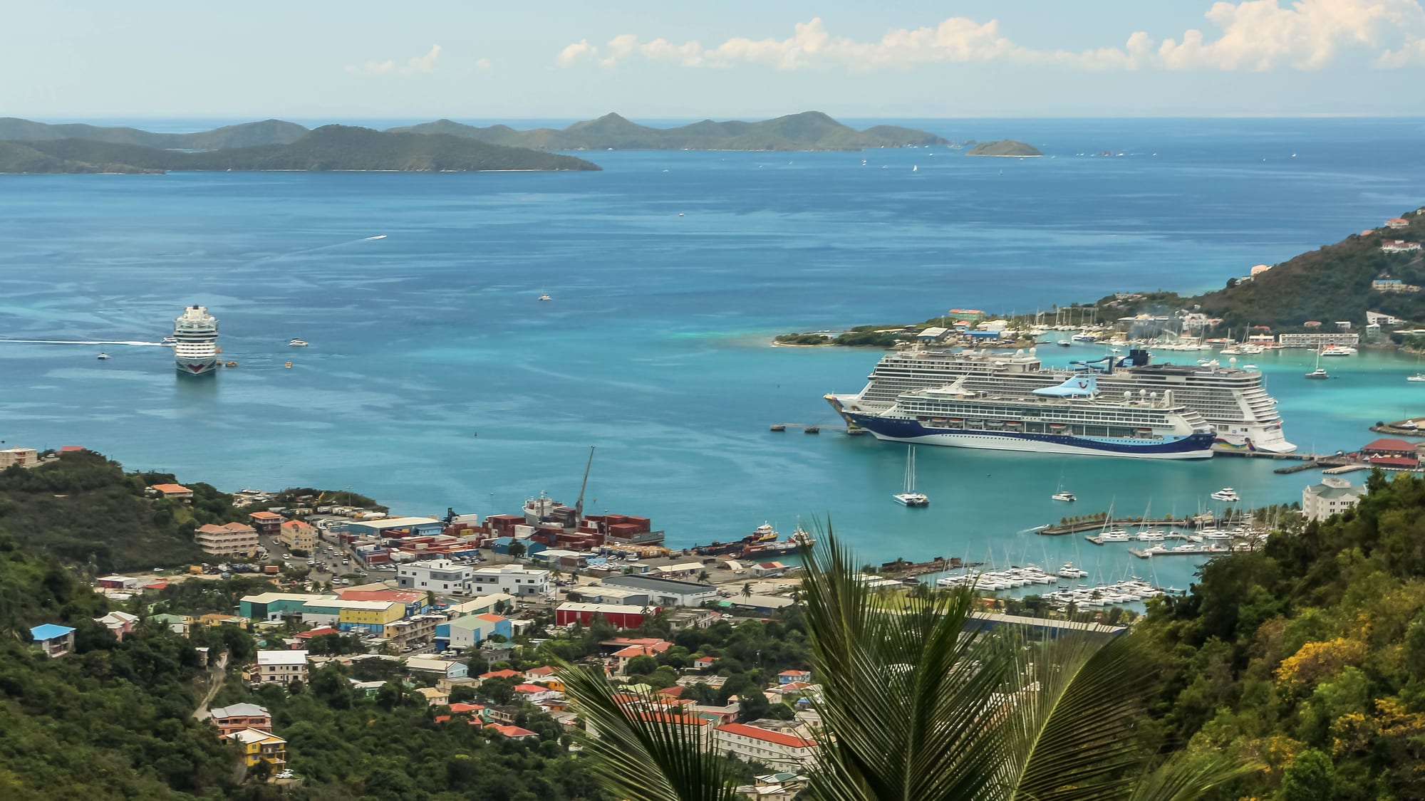 A green hilly coastline with a small town and marina along a sheltered blue bay dotted with boats