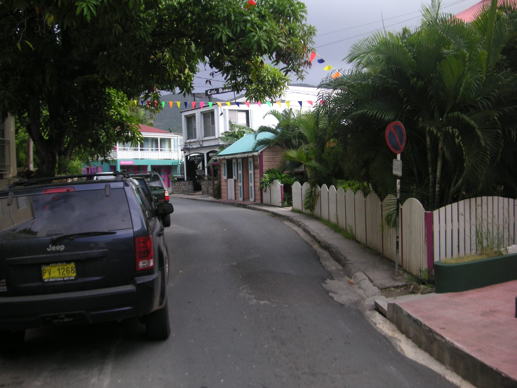 A narrow street curving past small painted wooden buildings, a white picket fence and palm trees, with strings of coloured pennants hung overhead and cars parked along one side