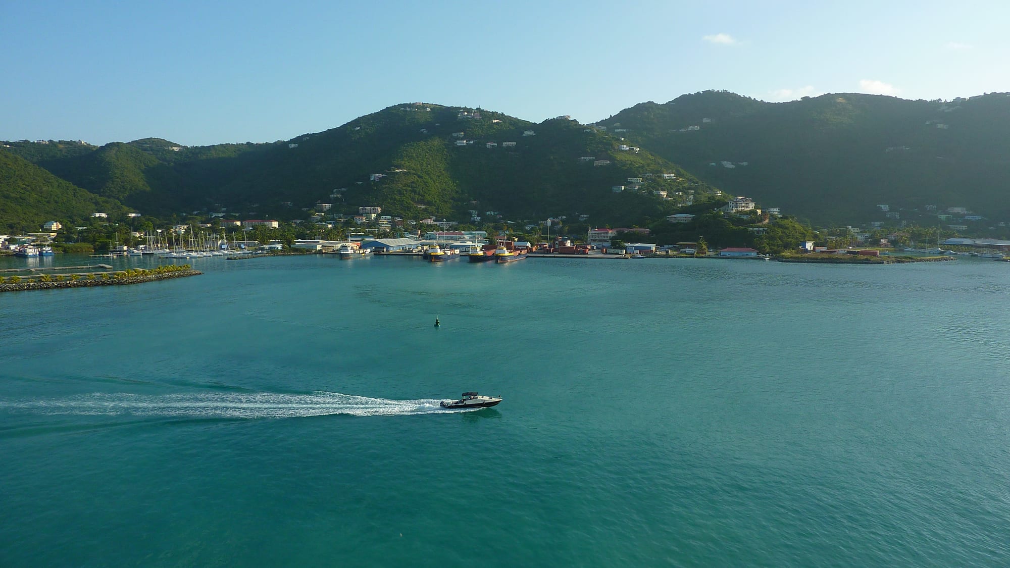 A wide turquoise harbour with a speedboat trailing white wake in the foreground, yachts moored at a marina on the left and a town spread along the shore beneath green hills