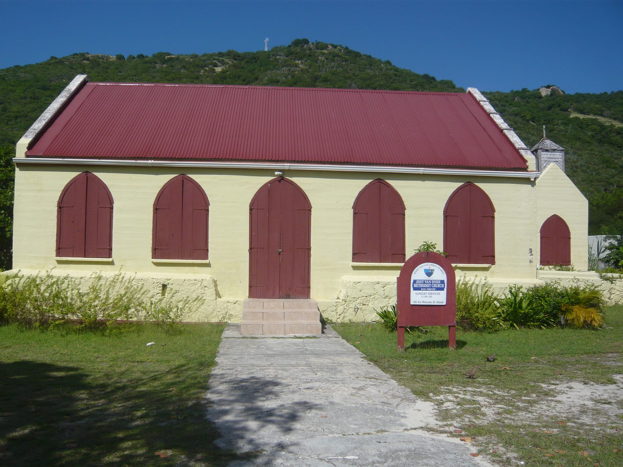 A small pale yellow church with a red corrugated roof, red shuttered arched windows and doors, and a noticeboard on the lawn, set against a green hillside