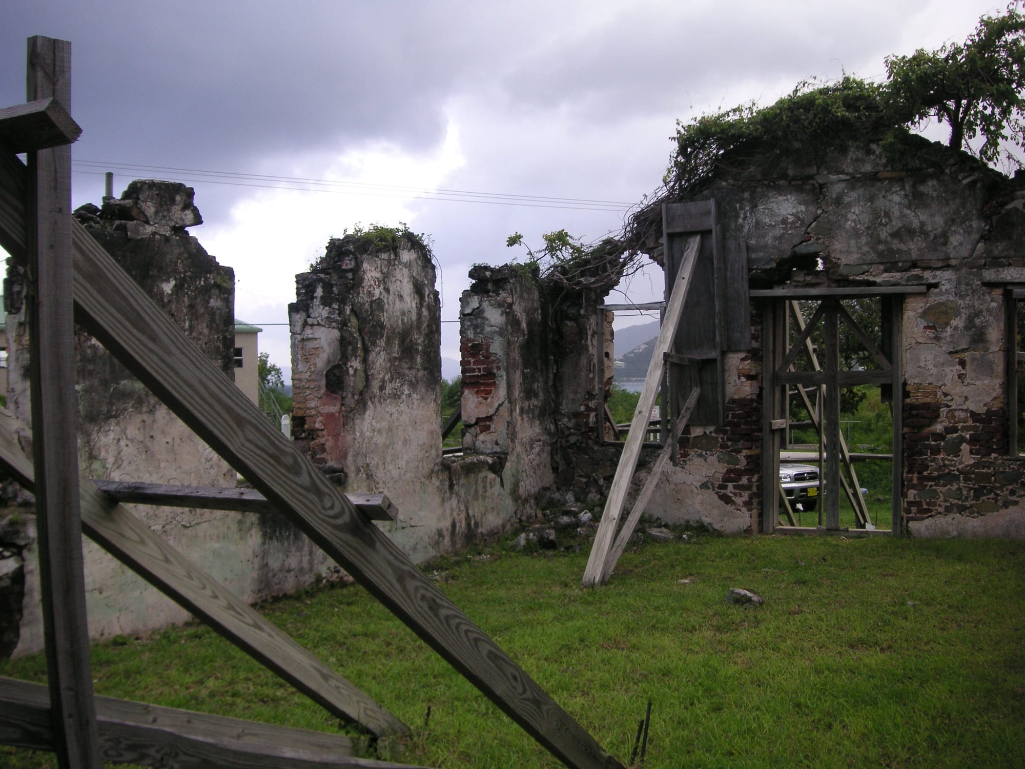 The roofless stone and brick walls of a ruined church overgrown with plants, propped up by diagonal timber supports, under a grey sky
