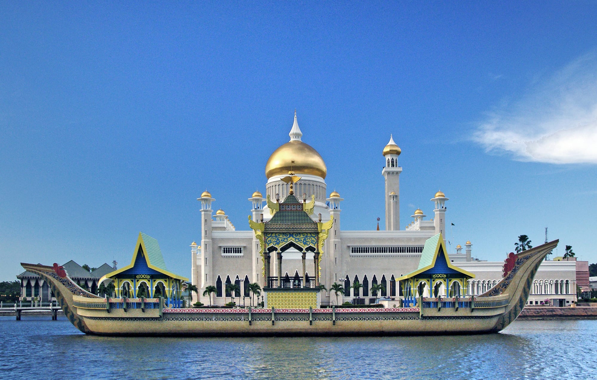 A white mosque with a large gold dome and a slender minaret, mirrored in a still lagoon with a stone barge