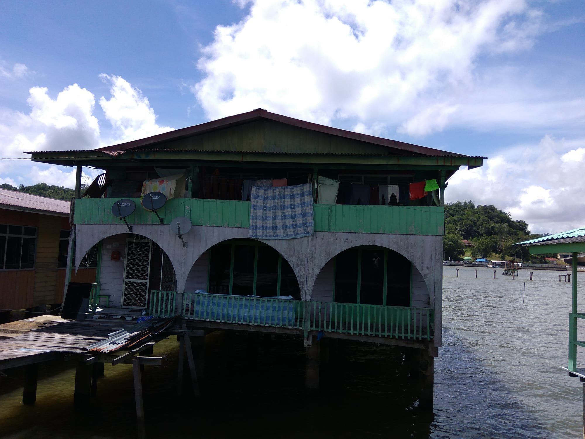 A two-storey timber house on stilts over the water, painted green and white with arched openings, laundry and a blanket hanging from its upper veranda, and a wooden walkway leading to it