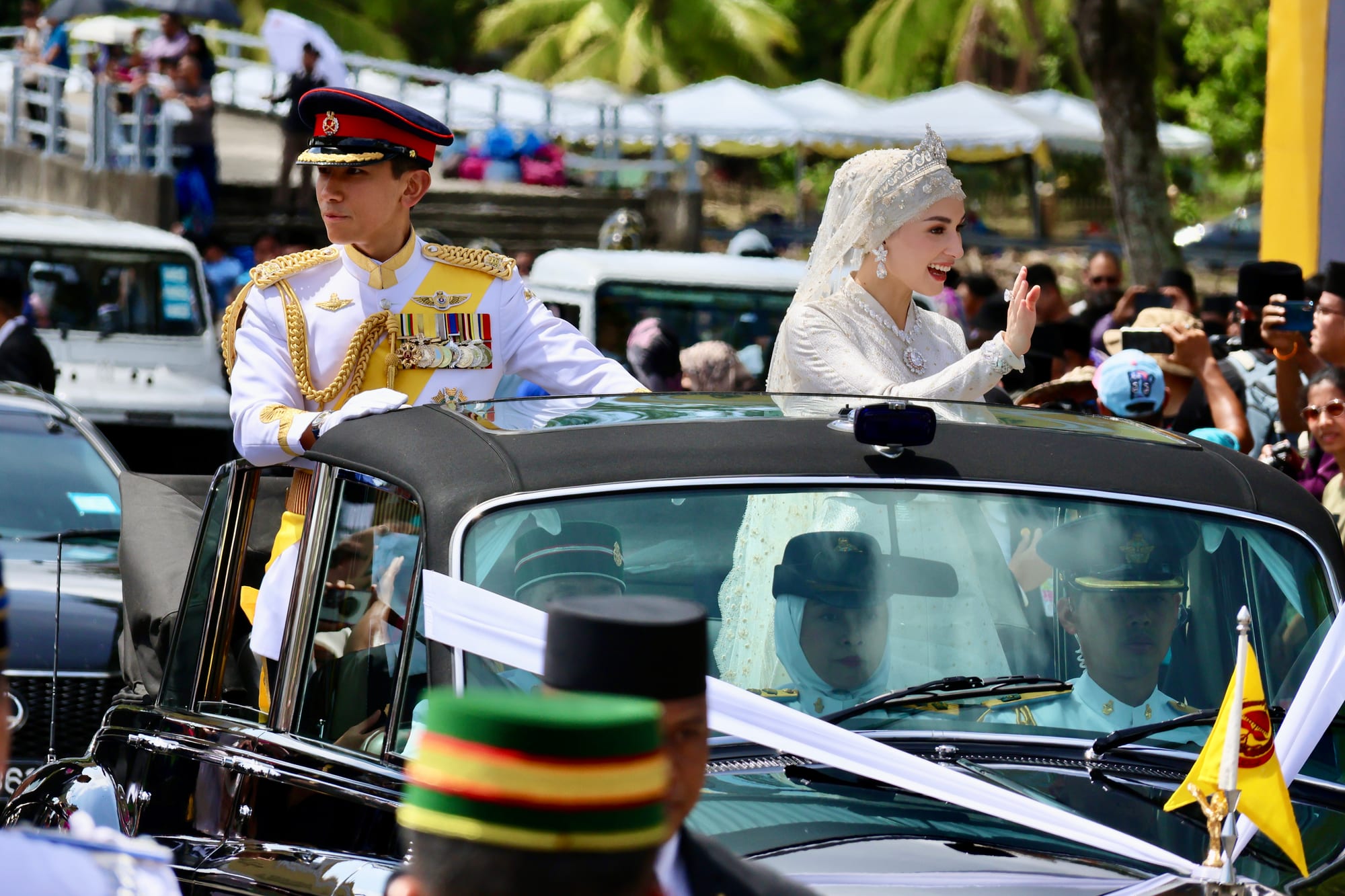 A groom in a white military dress uniform and a bride in a white embroidered veil and gown stand in an open-topped car, the bride waving, as crowds line the road behind them