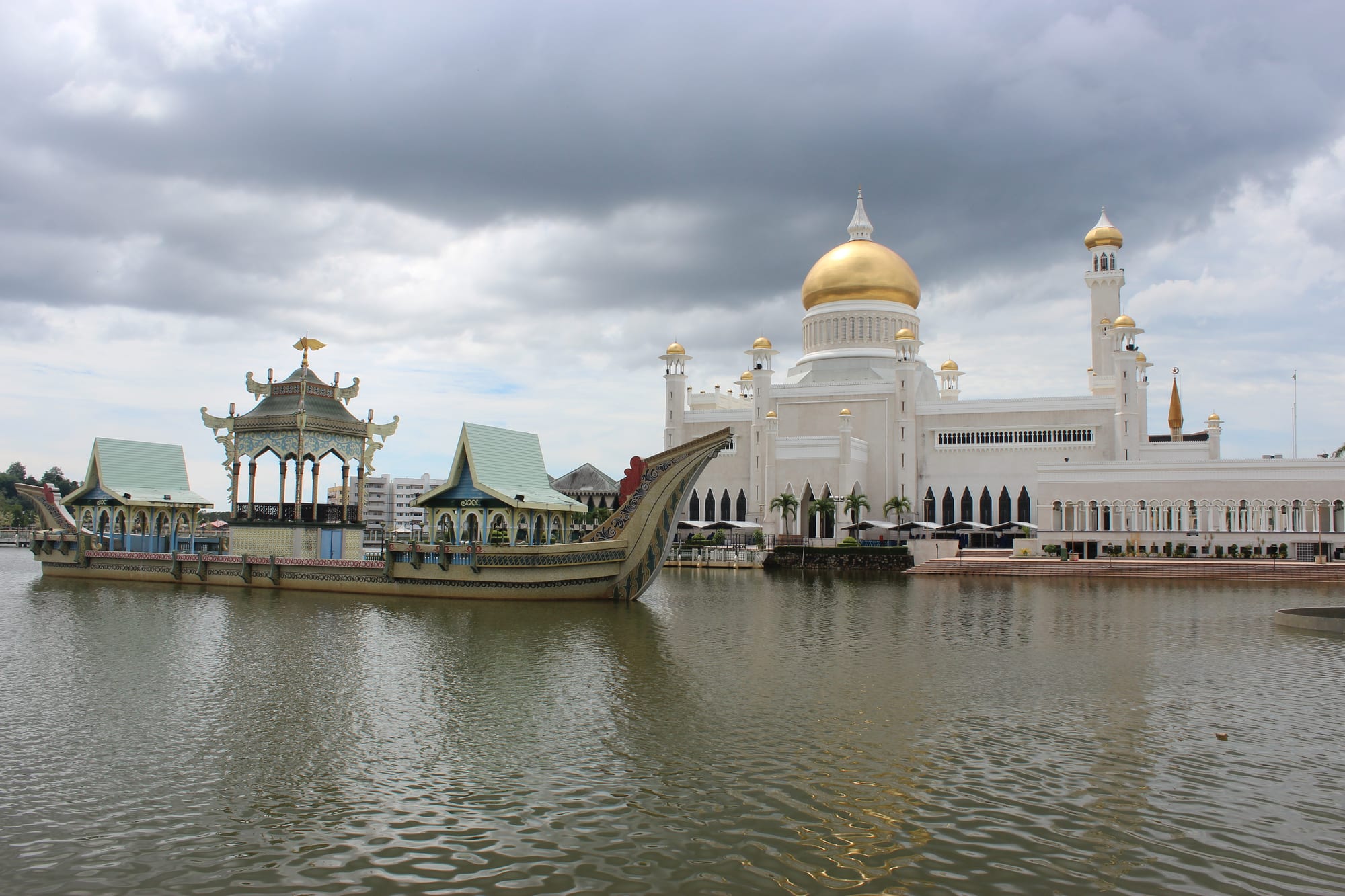 A white mosque with a large golden dome and slender minarets beside a lagoon, with an ornate barge topped by pavilions moored in the water in front