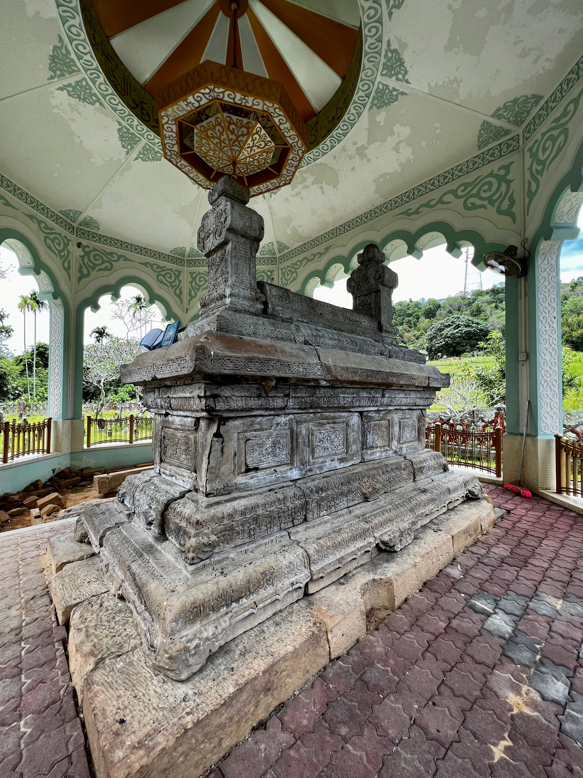 A weathered carved stone grave marker under a simple shelter, surrounded by other old tombs