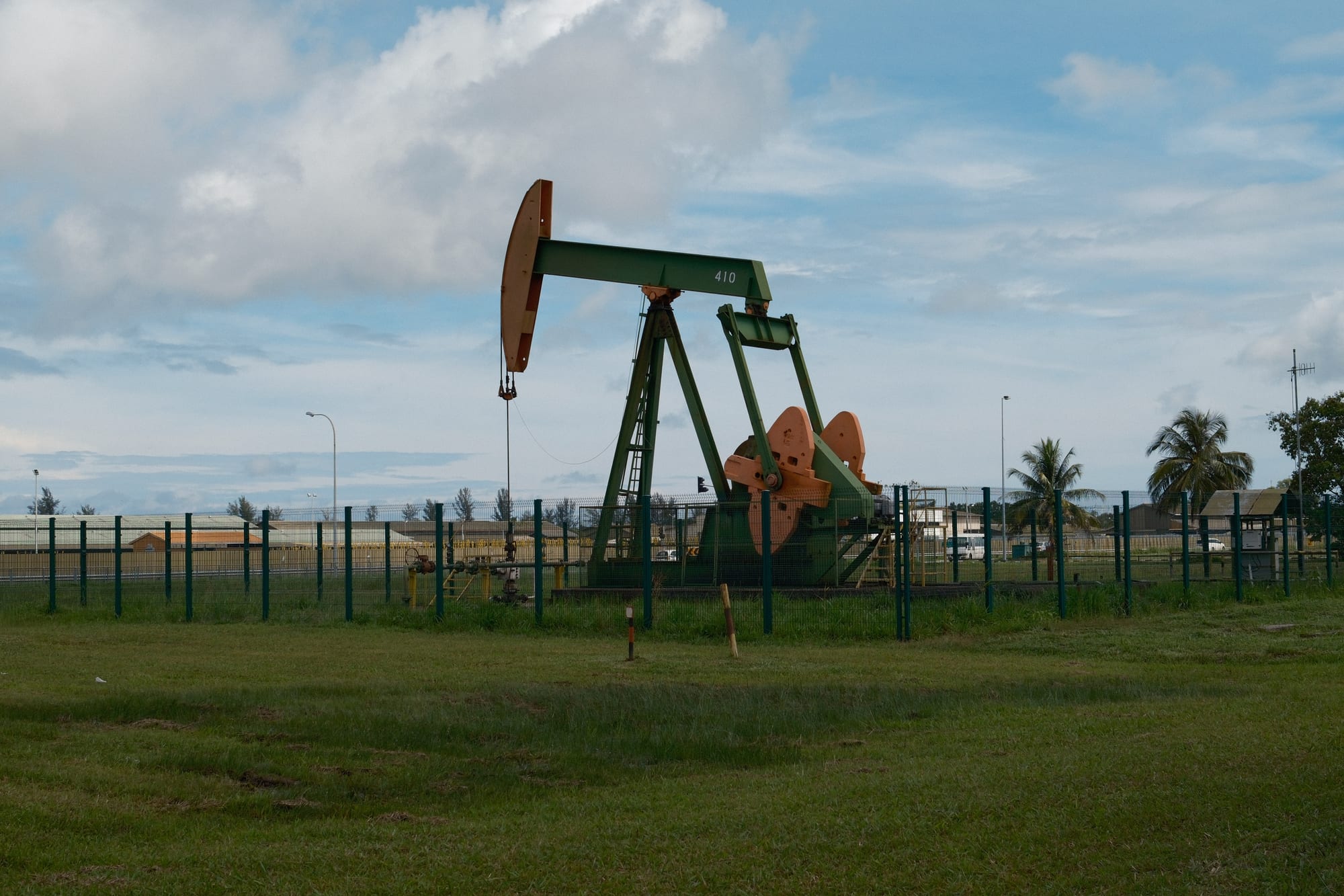 A green and orange nodding-donkey oil pump behind a fence on a grassy field, with palm trees and a cloudy sky