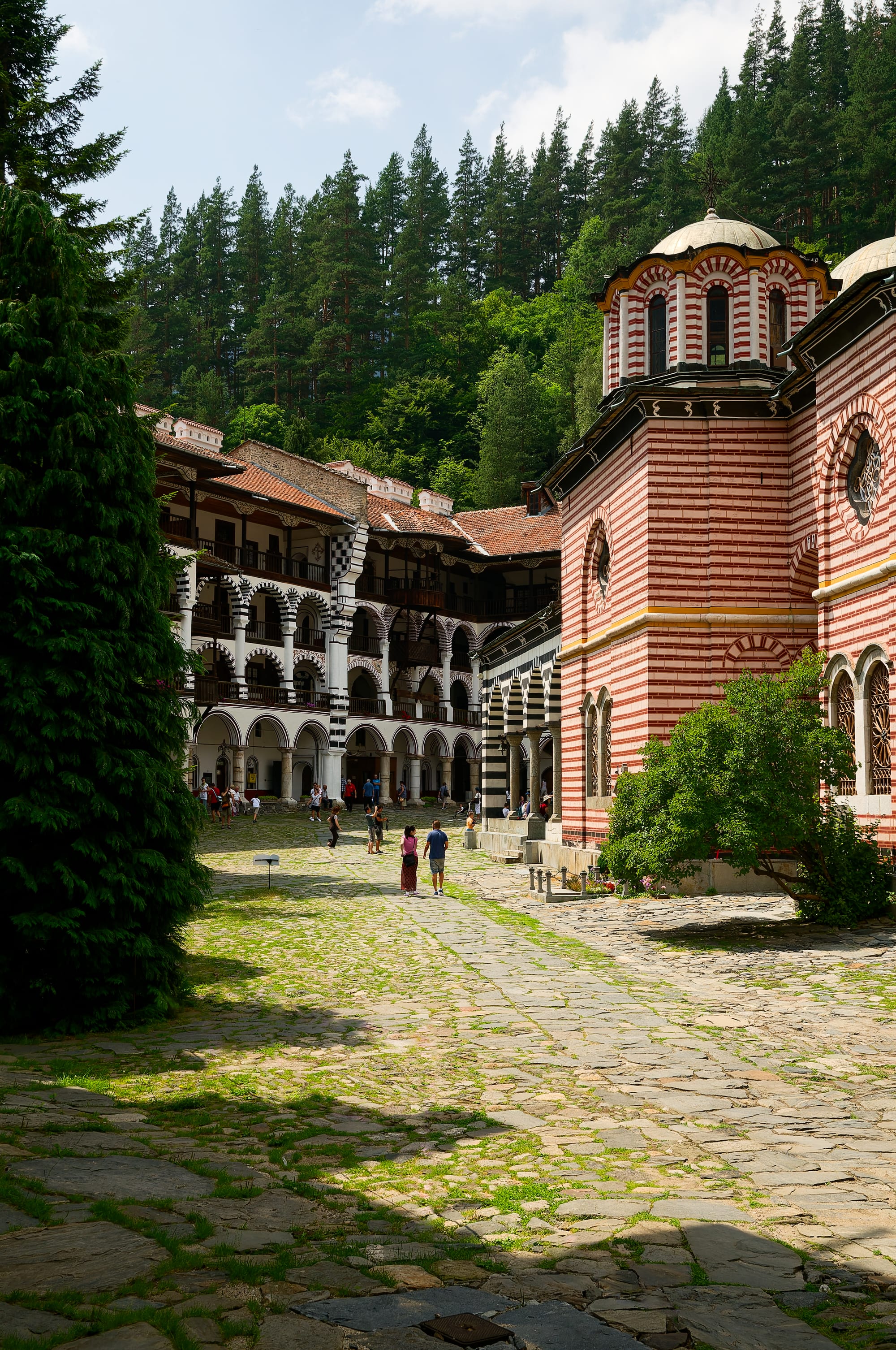 A tall striped monastery church with domes and a frescoed porch, mountains behind