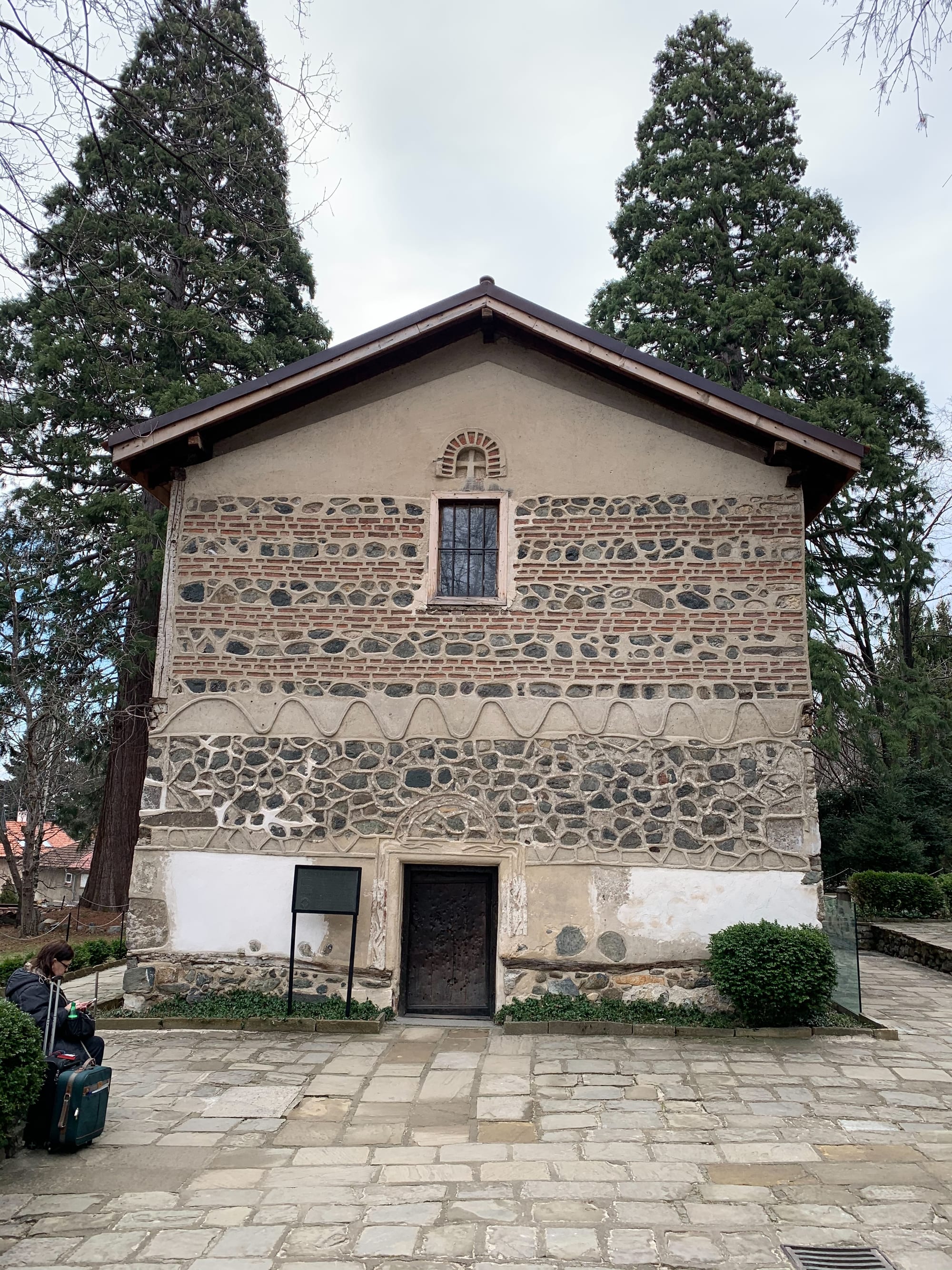 A small stone medieval church with a tiled roof, half-hidden among tall trees