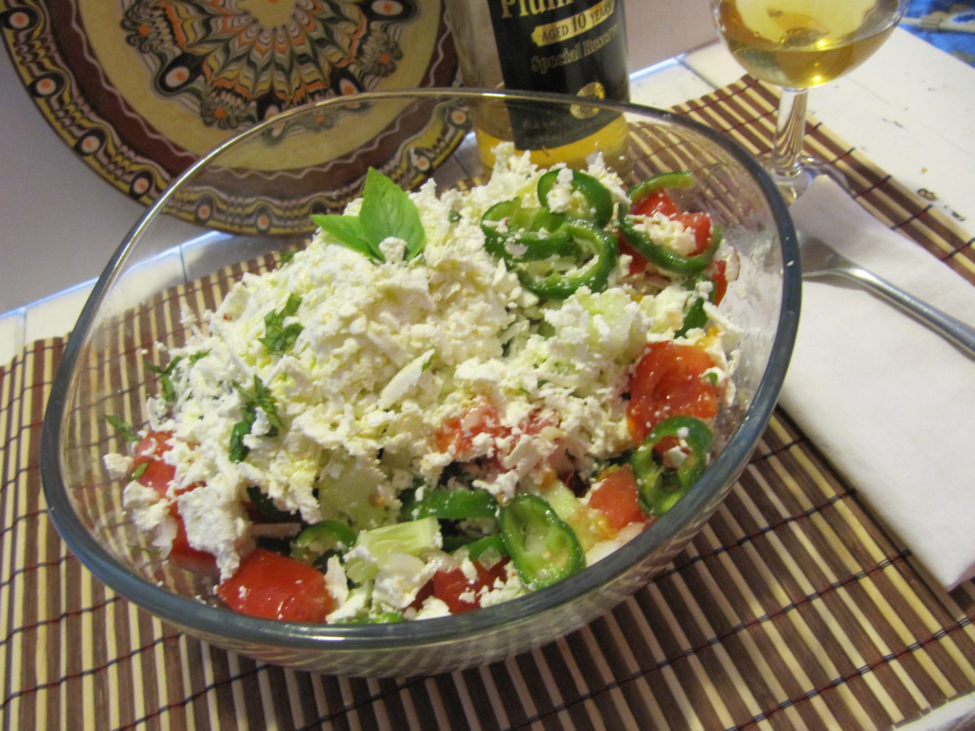 A bowl of chopped tomato, cucumber and pepper topped with grated white cheese, beside a small glass of clear brandy