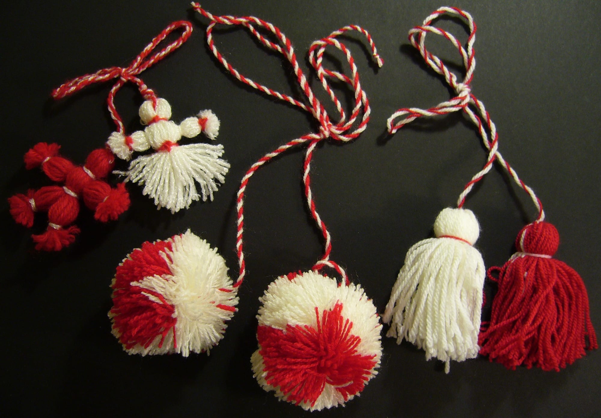 Several handmade red and white woollen tokens on a black background, including two small tasselled dolls, two pompoms and a pair of tassels, each on a twisted red and white cord
