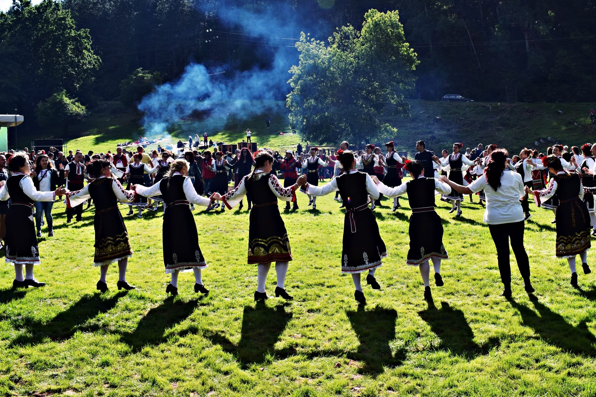 A long line of women in dark embroidered folk dresses and white blouses dance hand in hand across a sunlit meadow, with a large crowd and a wooded hillside behind