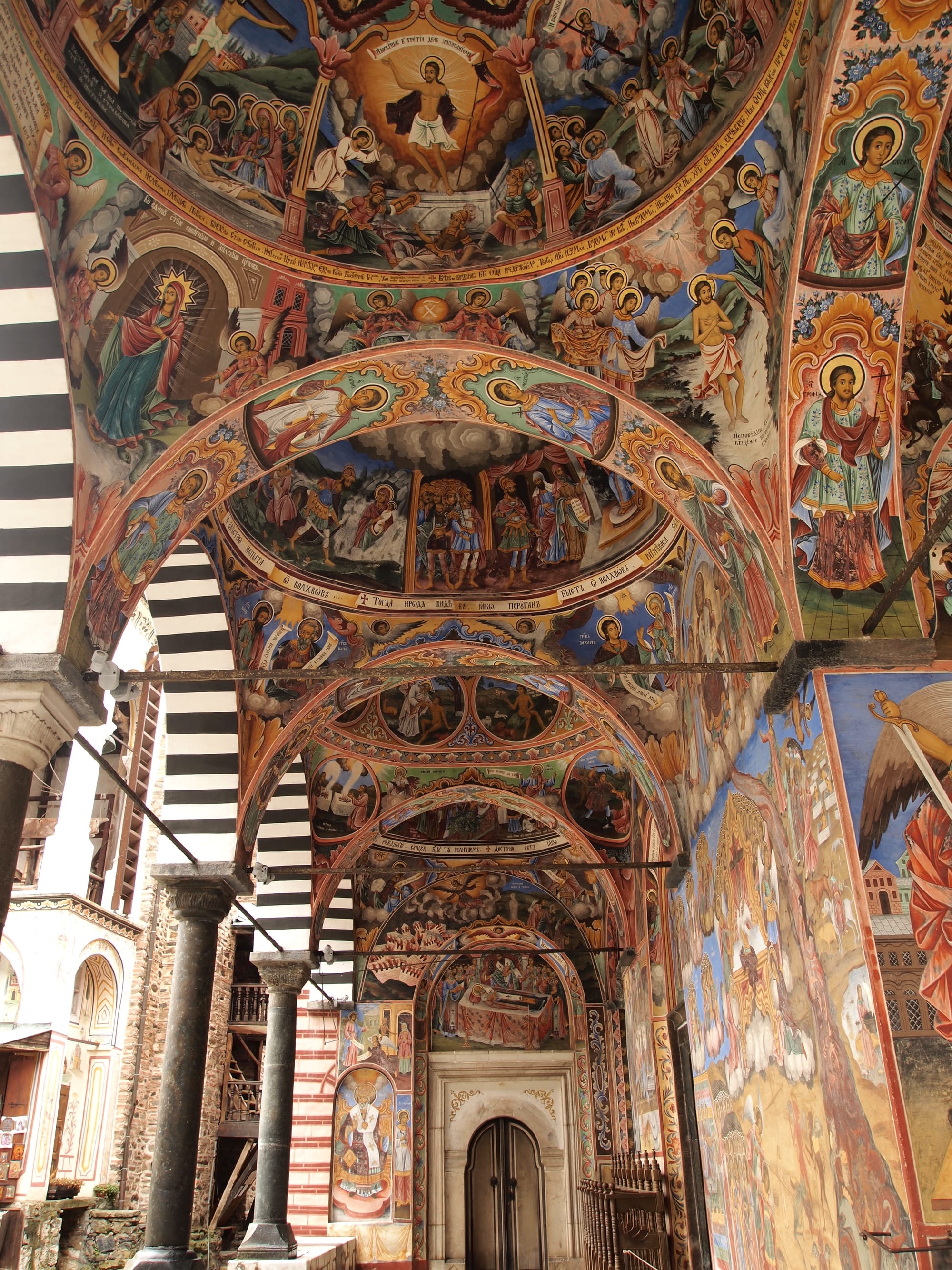 A vaulted arcade with striped black and white arches and columns, its ceiling and walls covered in brightly coloured religious paintings of saints and biblical scenes