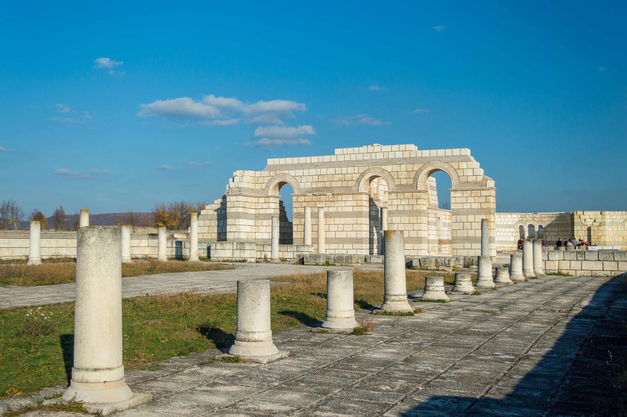 Rows of pale stone column stumps leading to a partly rebuilt basilica wall with three arches, under a deep blue sky