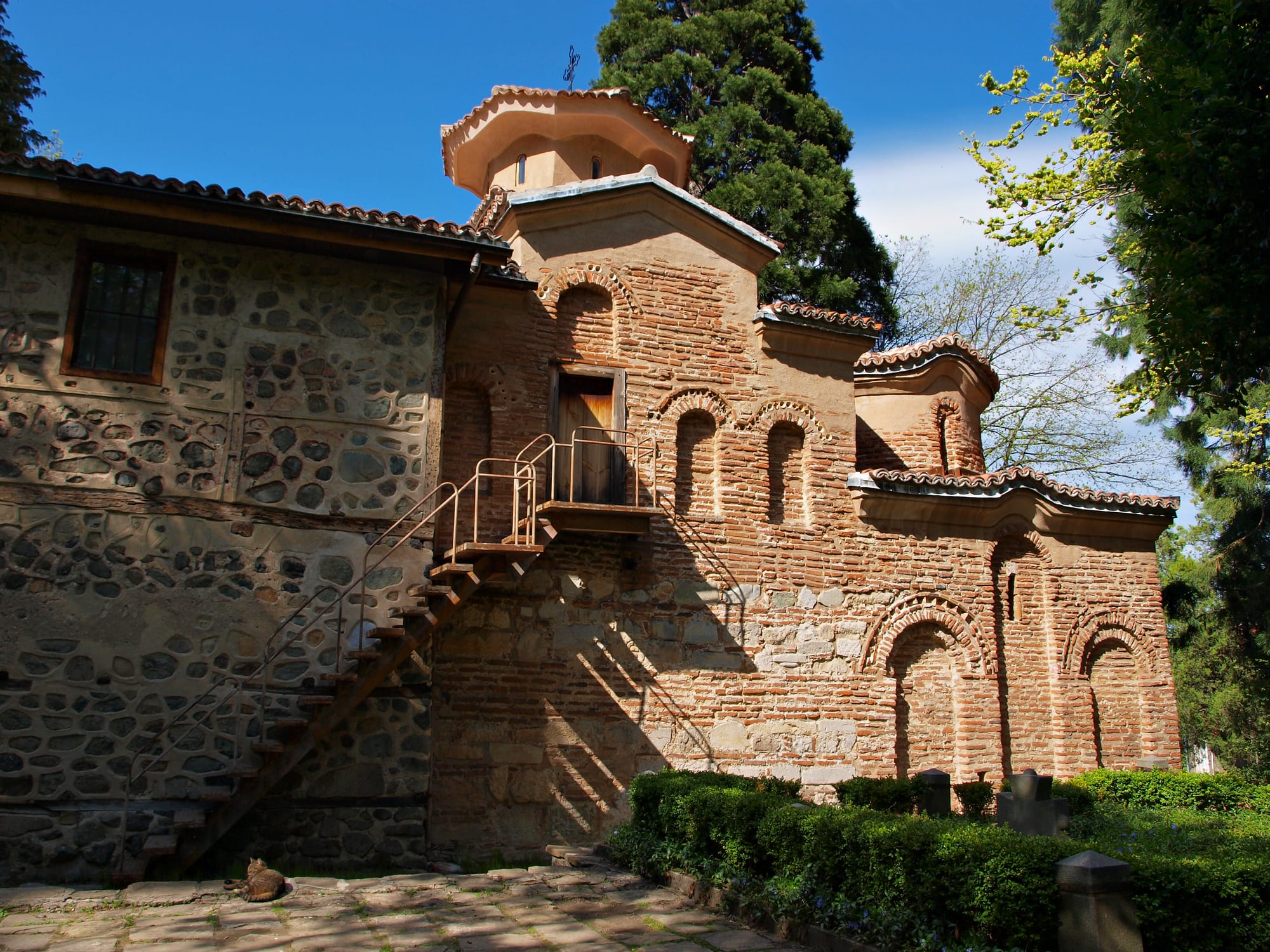 A small medieval stone church with a tiled roof and blind arcading, half-hidden among trees